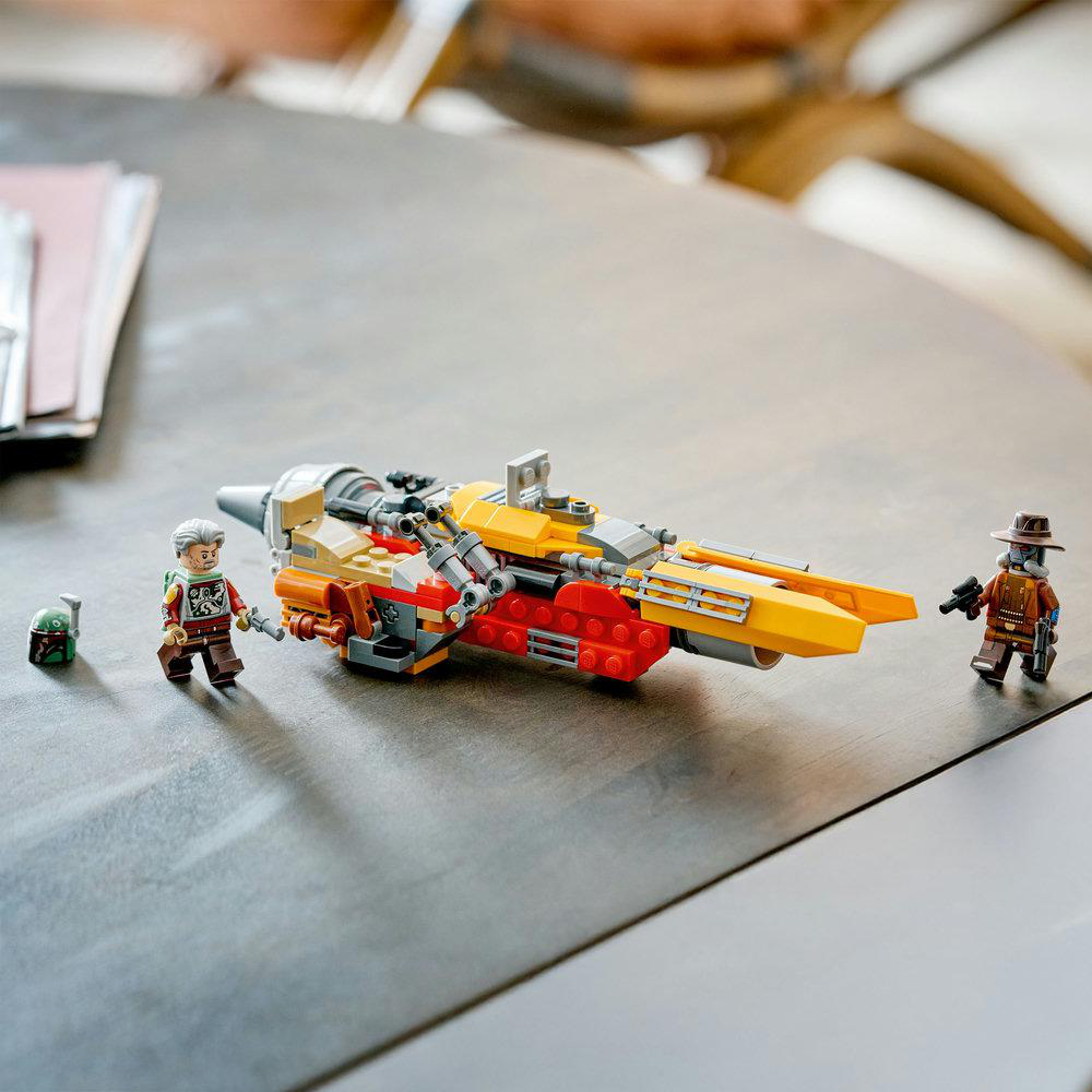 Ein Lego-Set mit einem gelb-roten Raumschiff und drei Minifiguren, darunter zwei Charaktere und ein Helm, auf einer grauen Oberfläche.