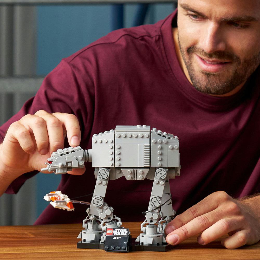 Ein Mann lächelt und hält ein Lego Star Wars AT-AT-Modell. Das Modell ist grau. Der Mann trägt ein weinrotes Hemd.