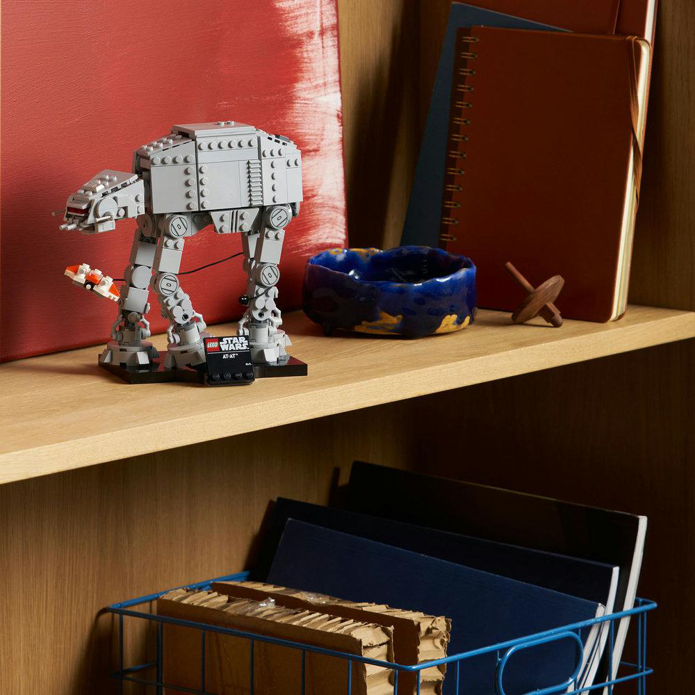 Ein Lego AT-AT-Modell auf einem Regal mit einer blauen Schale, Notizbüchern und einem Holzspielzeug. Im Hintergrund ein rotes Kunstwerk.