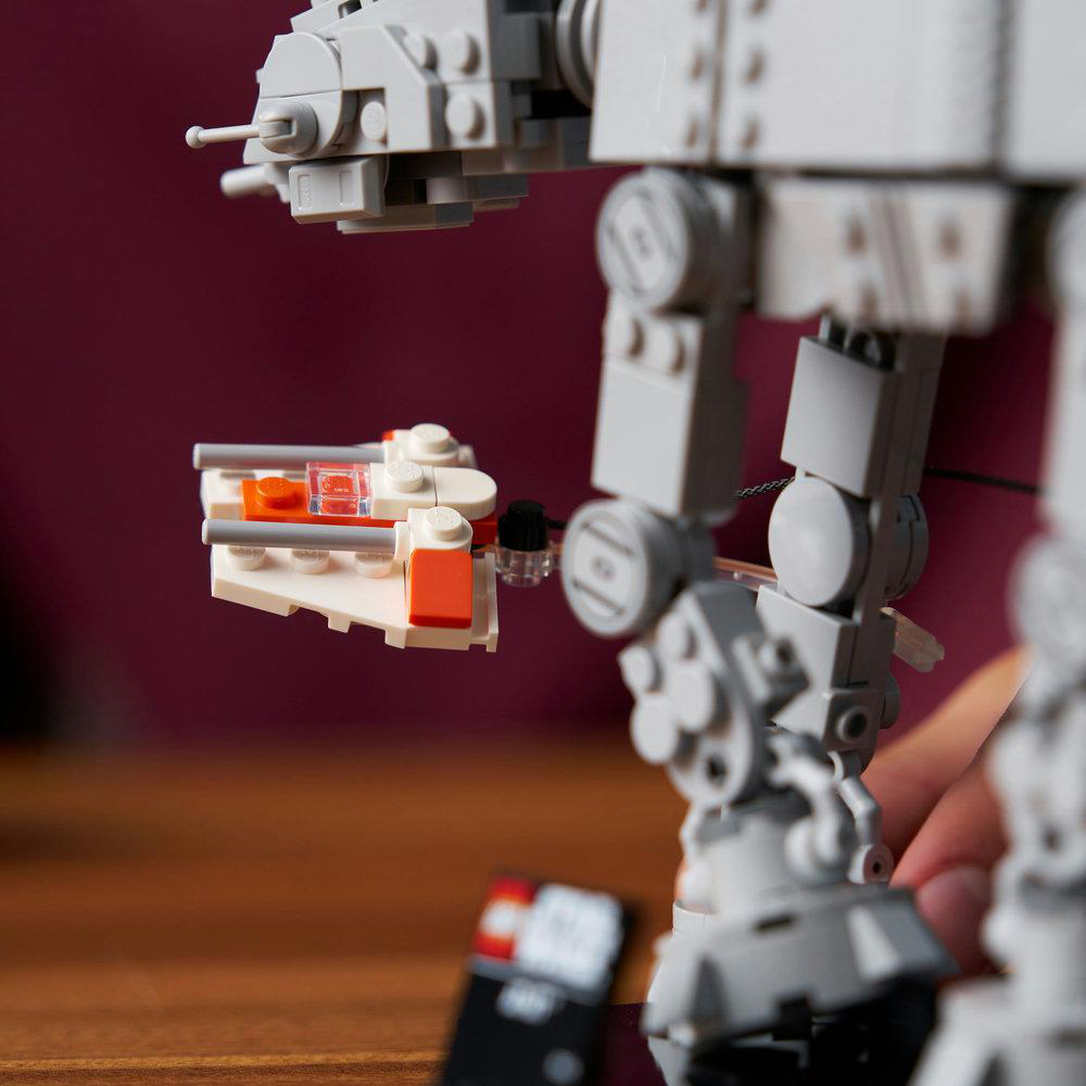 Ein Lego AT-AT Walker-Modell mit einem kleinen weißen und orangefarbenen Raumschiff. Der Hintergrund ist eine dunkelrote Oberfläche.