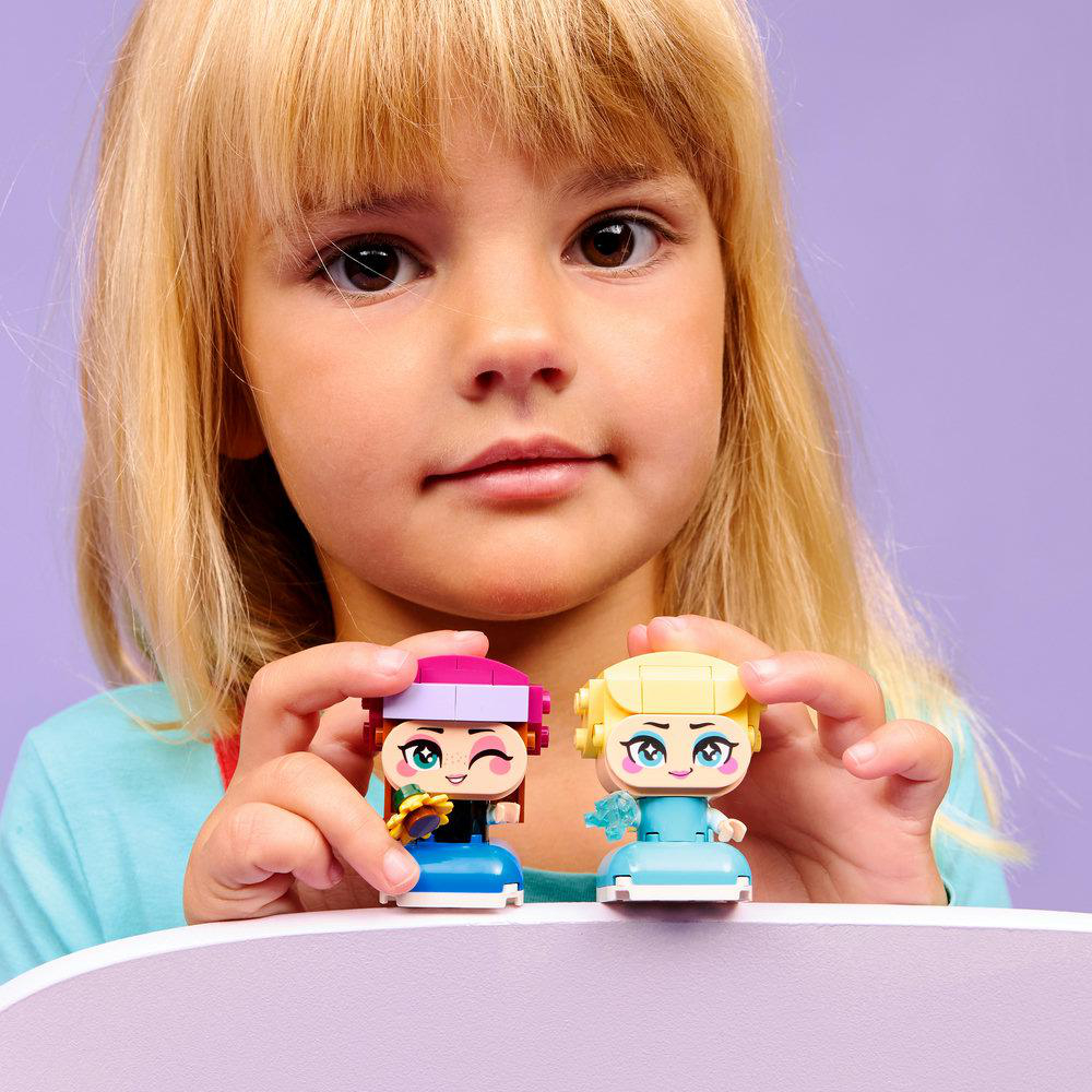 Ein Mädchen hält zwei Lego-Figuren. Eine hat rosa Haare und eine Blume, die andere hat gelbe Haare. Lila Hintergrund.
