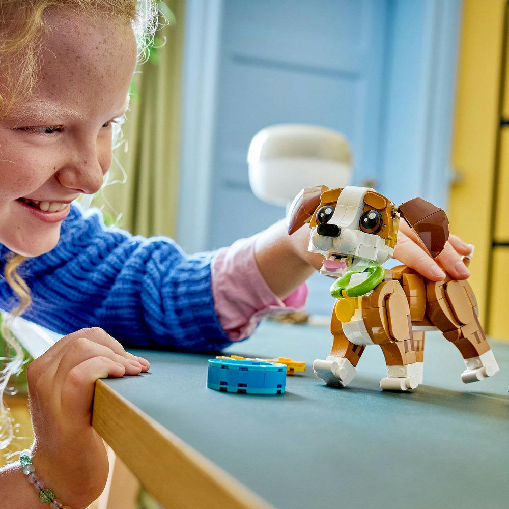 Ein Mädchen lächelt, während sie mit einem Lego-Hund interagiert. Der Hund ist braun, weiß und hellbraun. Ein blaues Lego liegt auf dem Tisch.