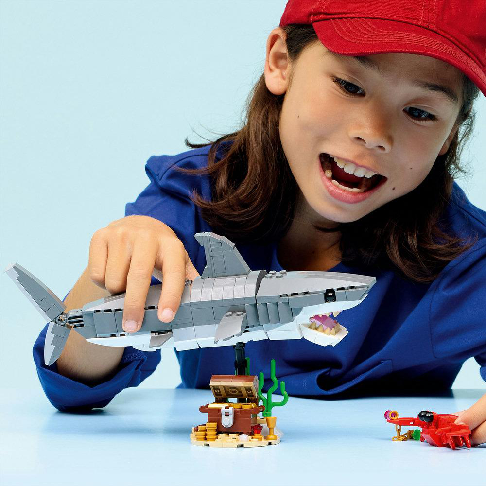 Ein Kind lächelt und hält einen Lego-Hai. Eine Schatzkiste und ein rotes Lego-Fahrzeug liegen auf einer blauen Oberfläche.