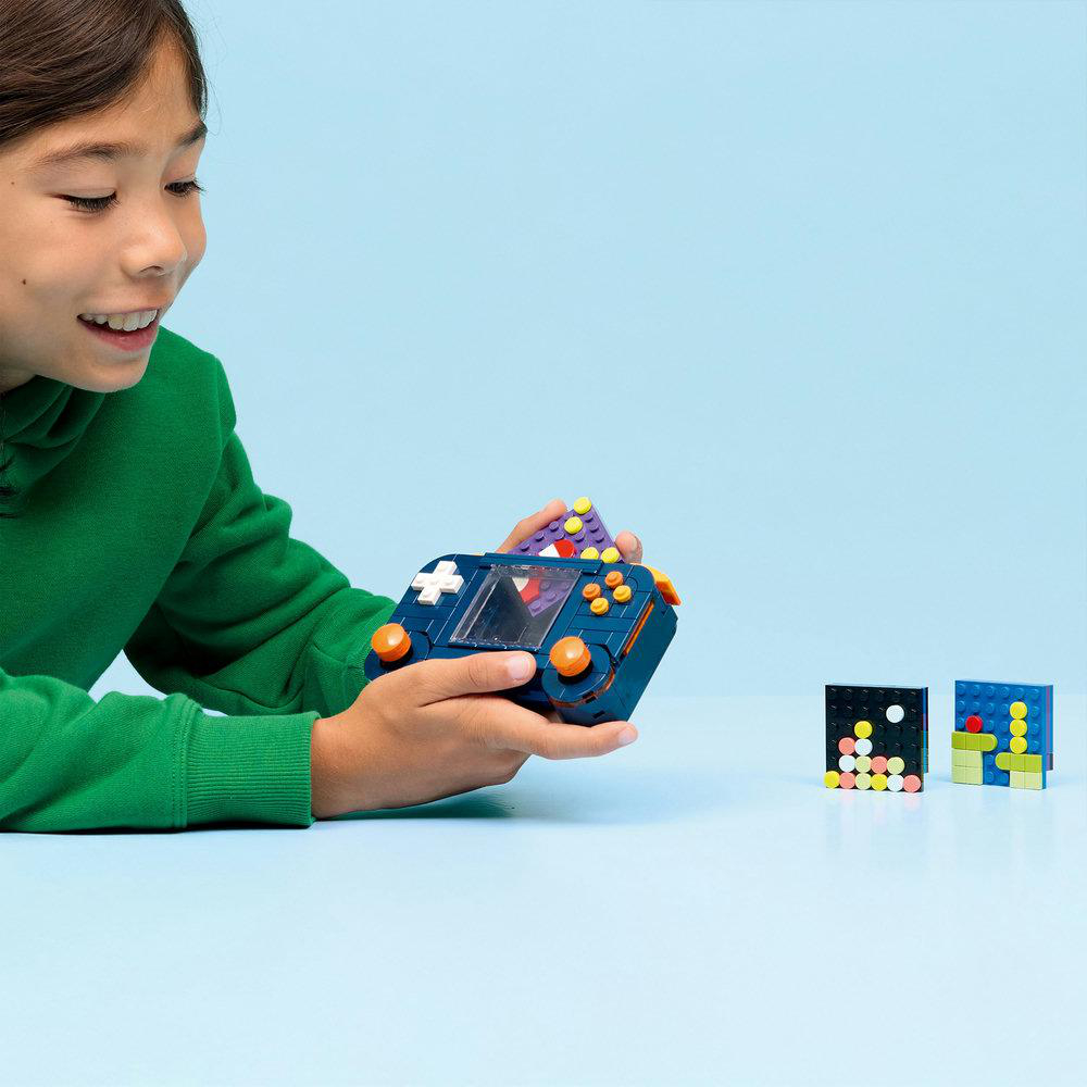 Ein Kind hält eine blaue Spielzeugkonsole mit orangefarbenen Knöpfen. Zwei pixelige Figuren sitzen auf dem Tisch, blauer Hintergrund.