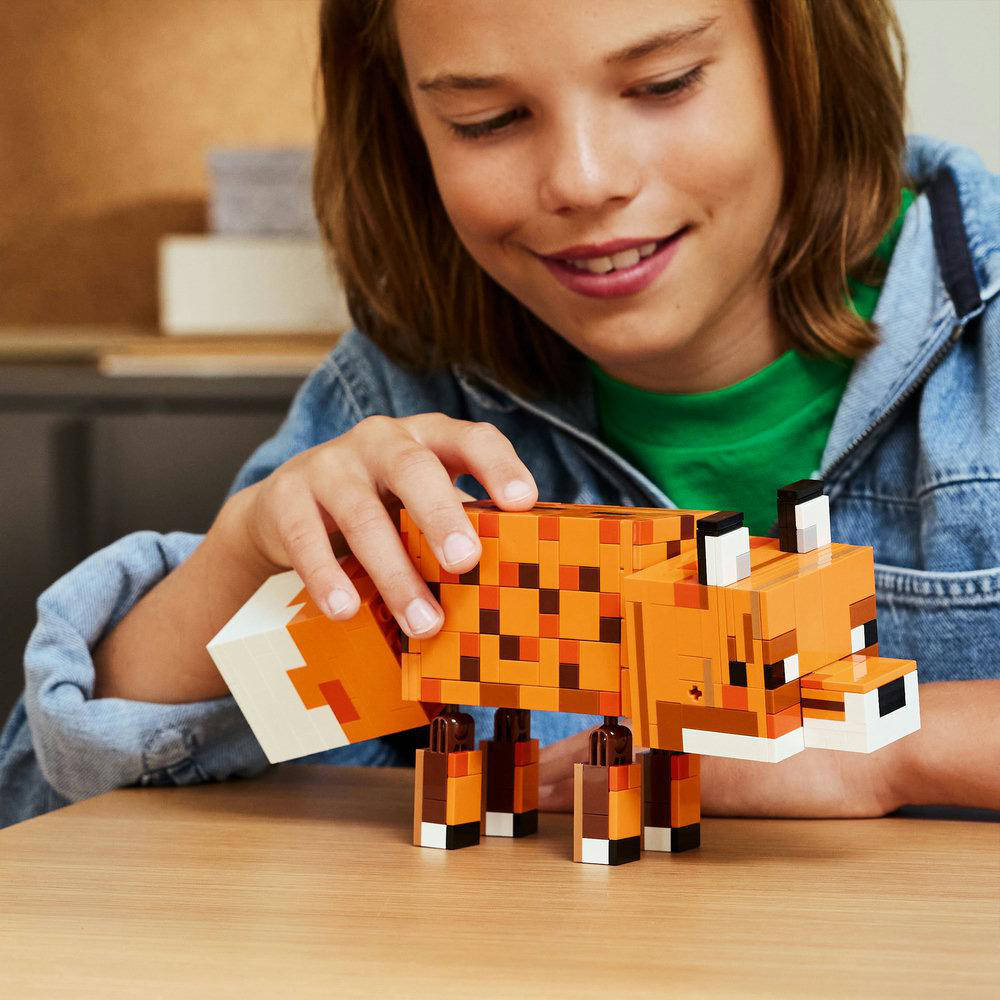Ein lächelnder Junge hält einen orangefarbenen Lego-Fuchs auf einem Holztisch. Der Fuchs hat braune Flecken, weiße und schwarze Details. Der Junge trägt eine Jeansjacke.