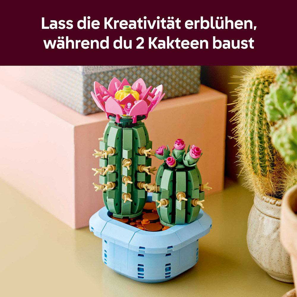 Zwei Lego-Kakteen mit rosa Blüten in einem blauen Topf. Hintergrund: grüner Kaktus und rosa Wand.