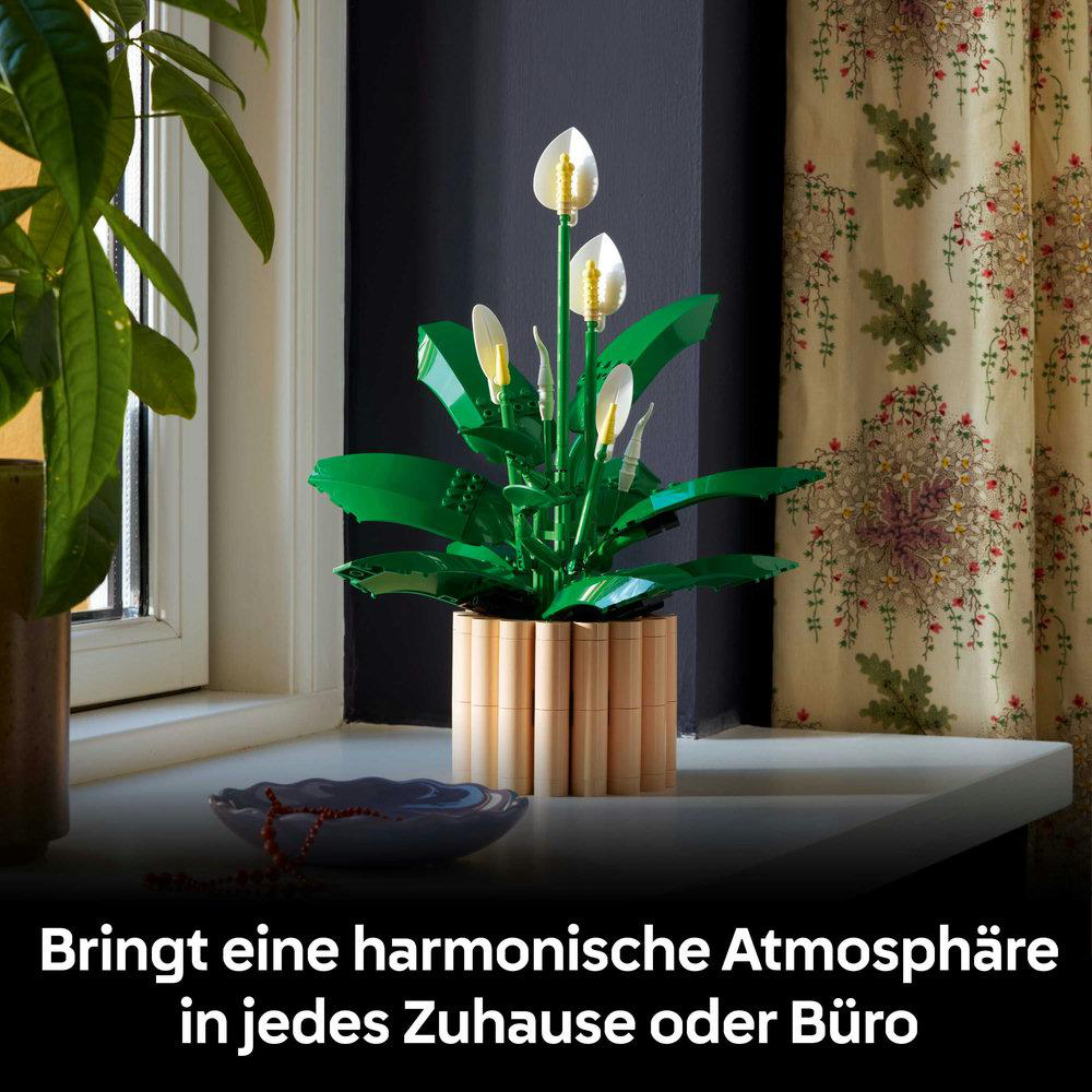 Eine Lego-Friedenslilie in einem Holztopf steht auf einer Fensterbank. Eine Schüssel und Perlen sind im Vordergrund. Im Hintergrund ein Vorhang.
