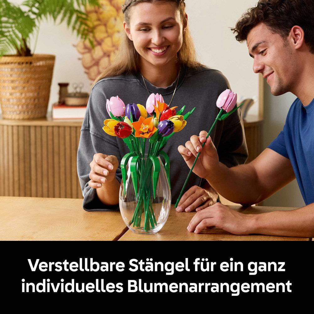 Ein Mann und eine Frau arrangieren bunte Lego-Blumen in einer Glasvase auf einem Holztisch. Sie lächeln.