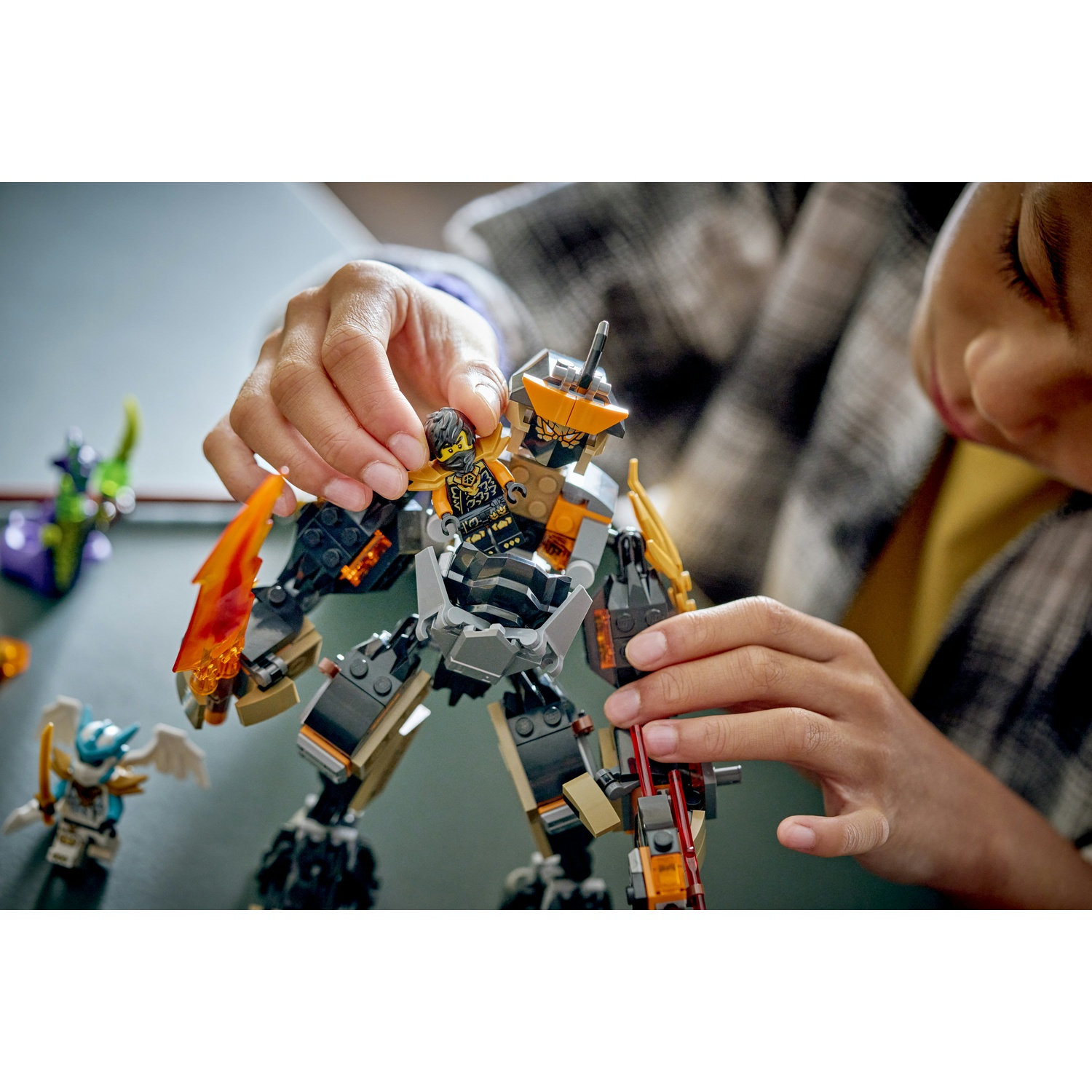 Una persona mette un minifigure in un mech Lego. Altre figure Lego sono sullo sfondo. Il mech è nero, grigio e arancione.