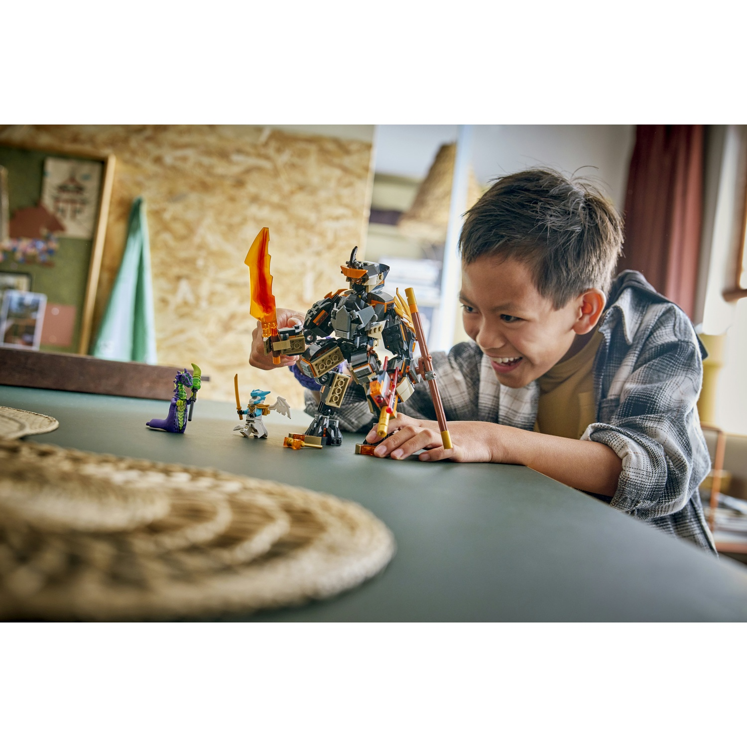 Un ragazzo sorride mentre gioca con i personaggi LEGO Ninjago su un tavolo. Una grande figura di robot con una spada infuocata è a fuoco.