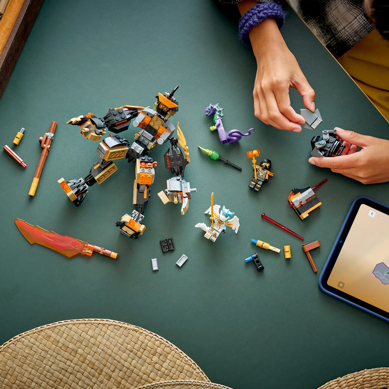 Un set Lego è sparso su una superficie verde. Le mani assemblano i pezzi. Sono visibili un robot, figurine e un tablet.