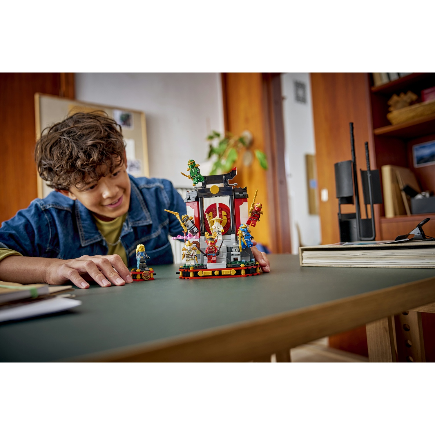 Un ragazzo sorride a un set Lego. Il set ha un edificio rosso e bianco con diversi personaggi Lego. Lo sfondo è una stanza.