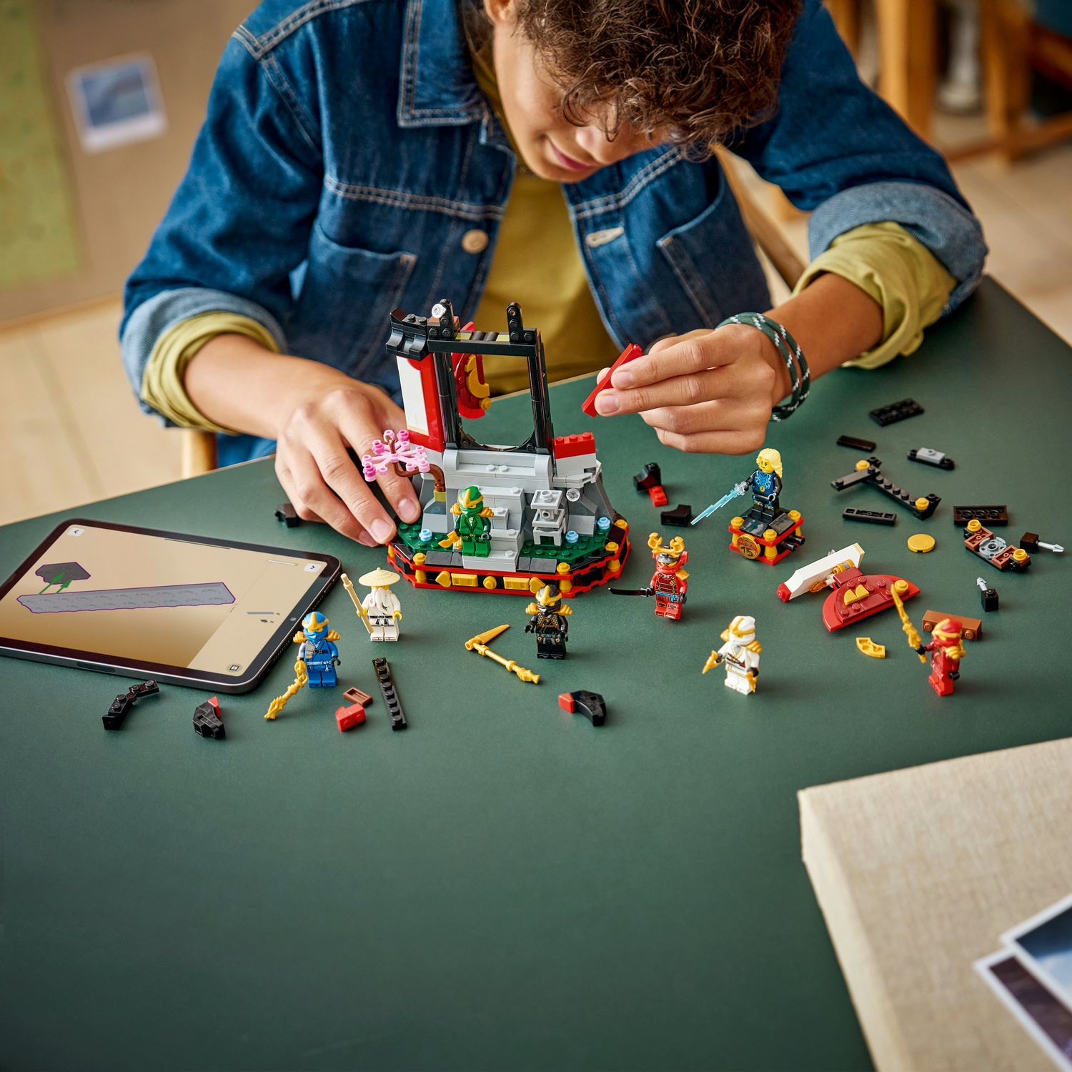 Un bambino sta assemblando un set Lego su un tavolo verde. Varie figure e pezzi Lego sono sparsi attorno a una struttura parzialmente costruita.
