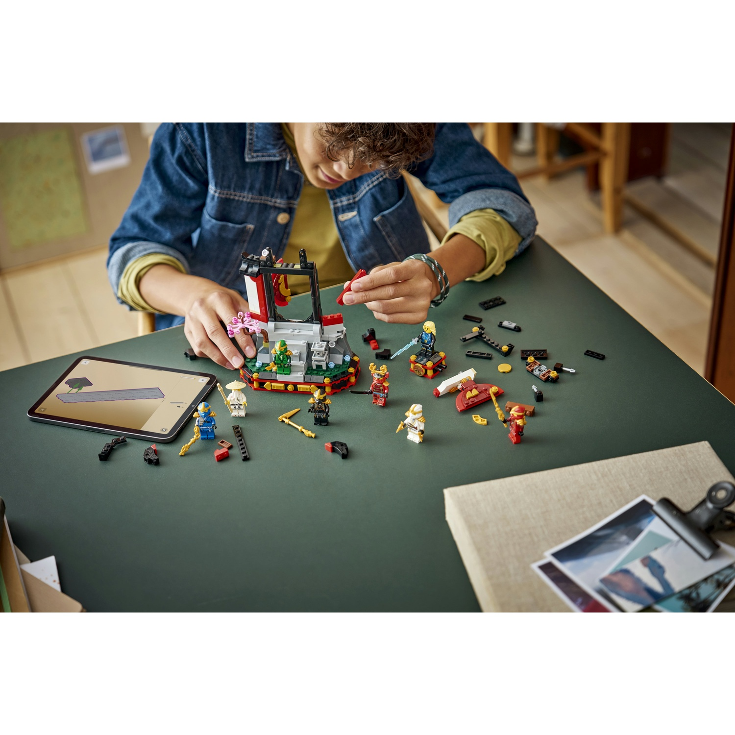 Una persona costruisce un set Lego su un tavolo verde. Il set include personaggi e strutture. Un tablet mostra le istruzioni. Altre parti sono sparse.