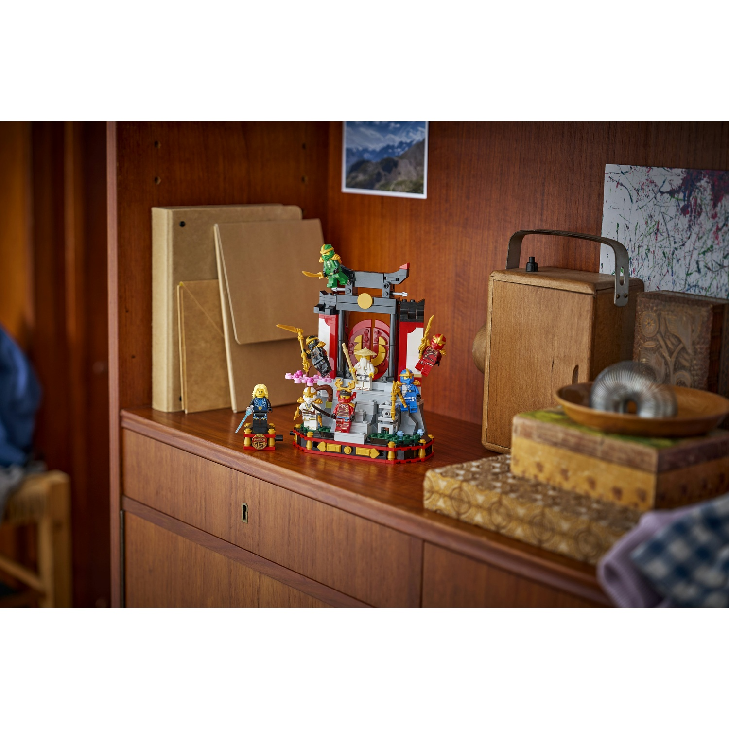Un set Lego è esposto su una mensola di legno. Il set raffigura una scena del tempio con diverse minifigure. Lo sfondo include libri e una scatola di legno.