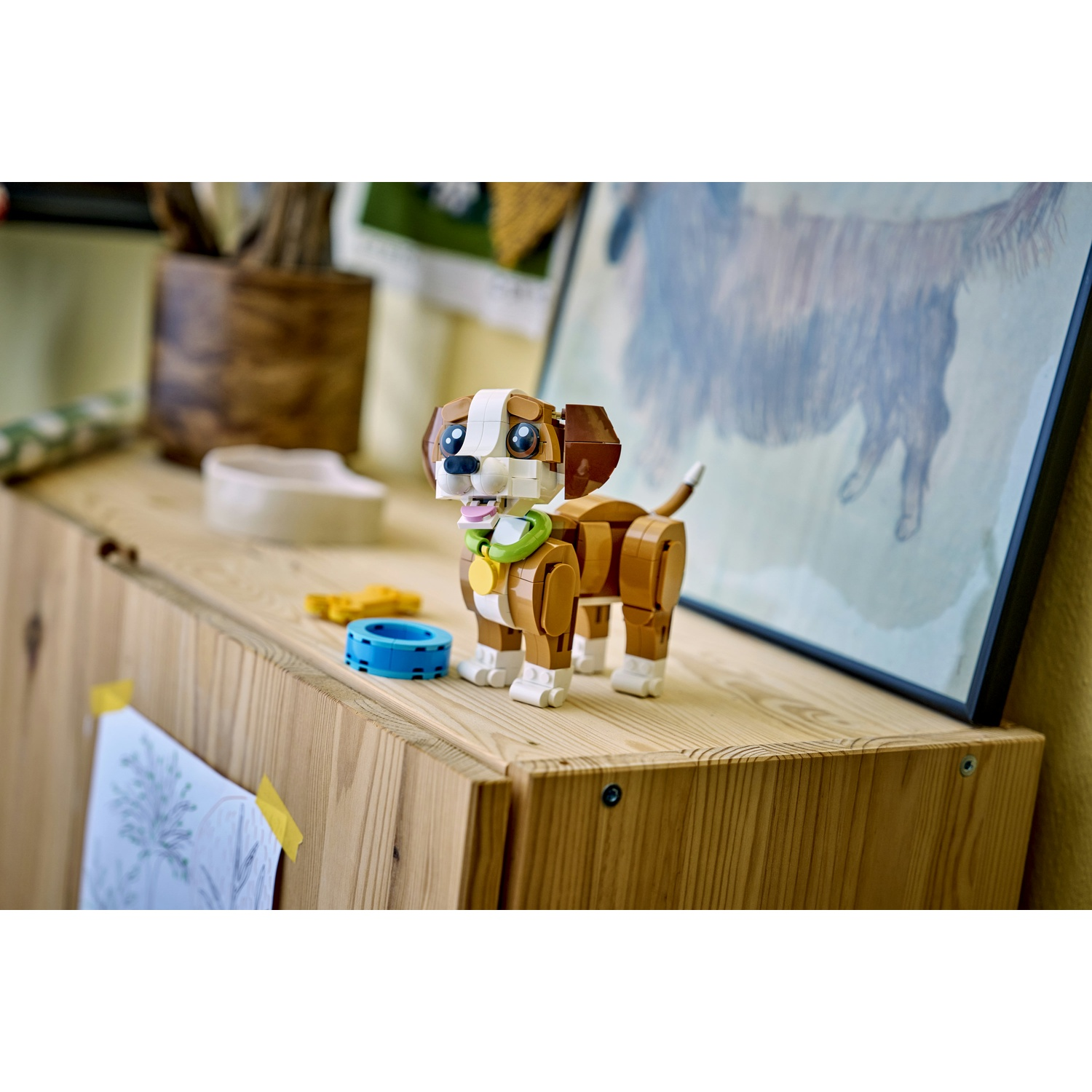 Un cane Lego è in piedi su uno scaffale di legno, con un anello blu e un osso giallo accanto. Sullo sfondo c'è un disegno di cane incorniciato.