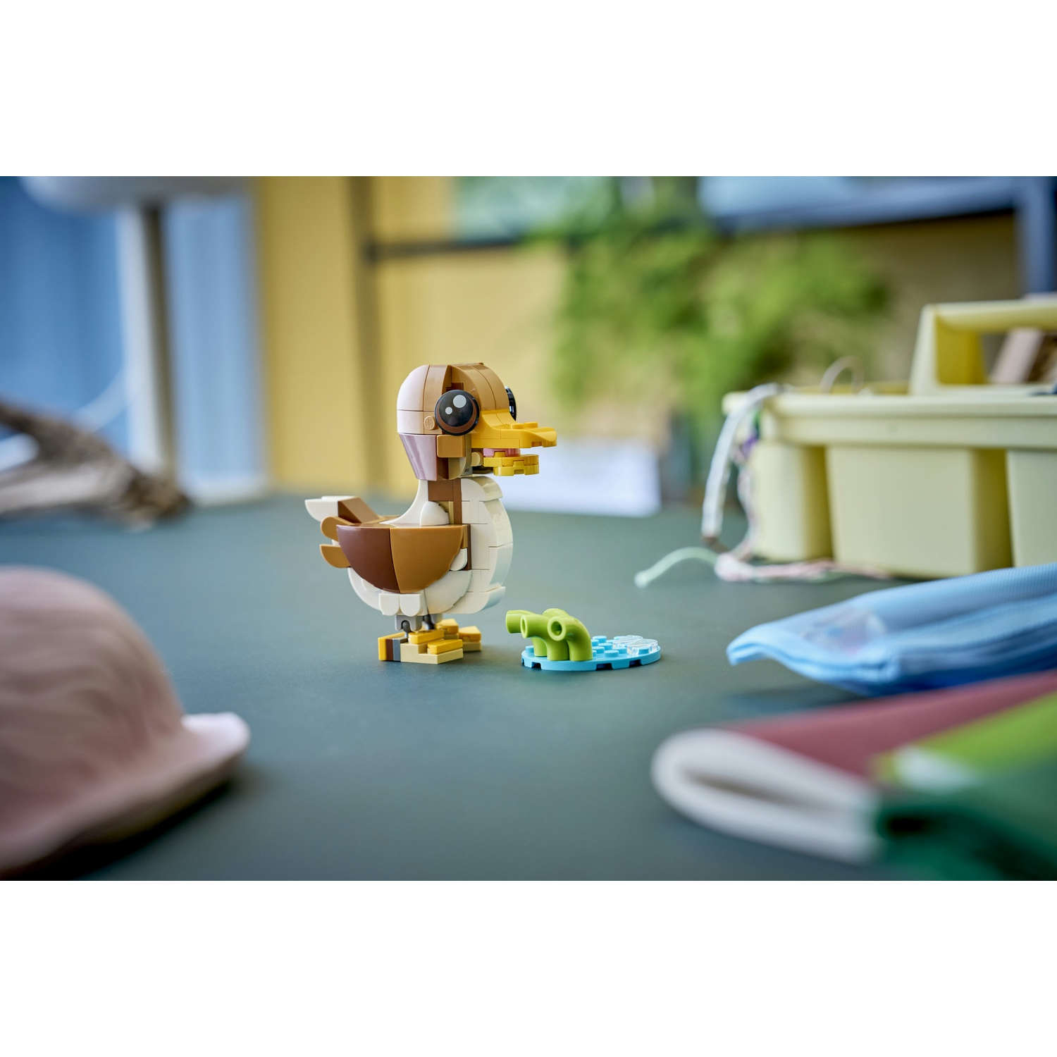 Un'anatra Lego, marrone e bianca, è su una superficie. Accanto, c'è un piccolo pezzo blu e verde. Altri oggetti sono sfocati sullo sfondo.