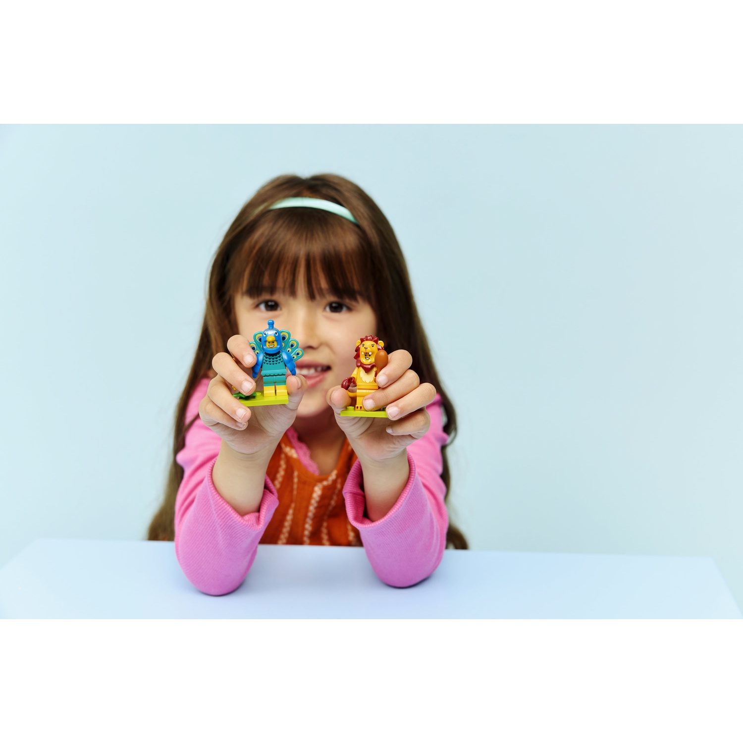 Una ragazza tiene in mano due personaggi Lego: un pavone e un leone. Indossa un top rosa e siede dietro un tavolo blu. Sfondo azzurro chiaro.