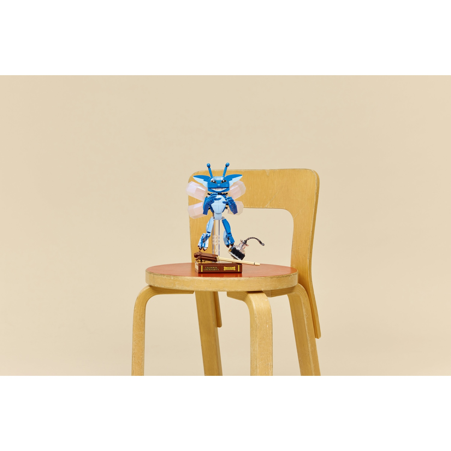 Una figura blu simile a un insetto è seduta su una sedia di legno. Un libro e un piccolo oggetto sono sulla sedia. Lo sfondo è una parete beige.