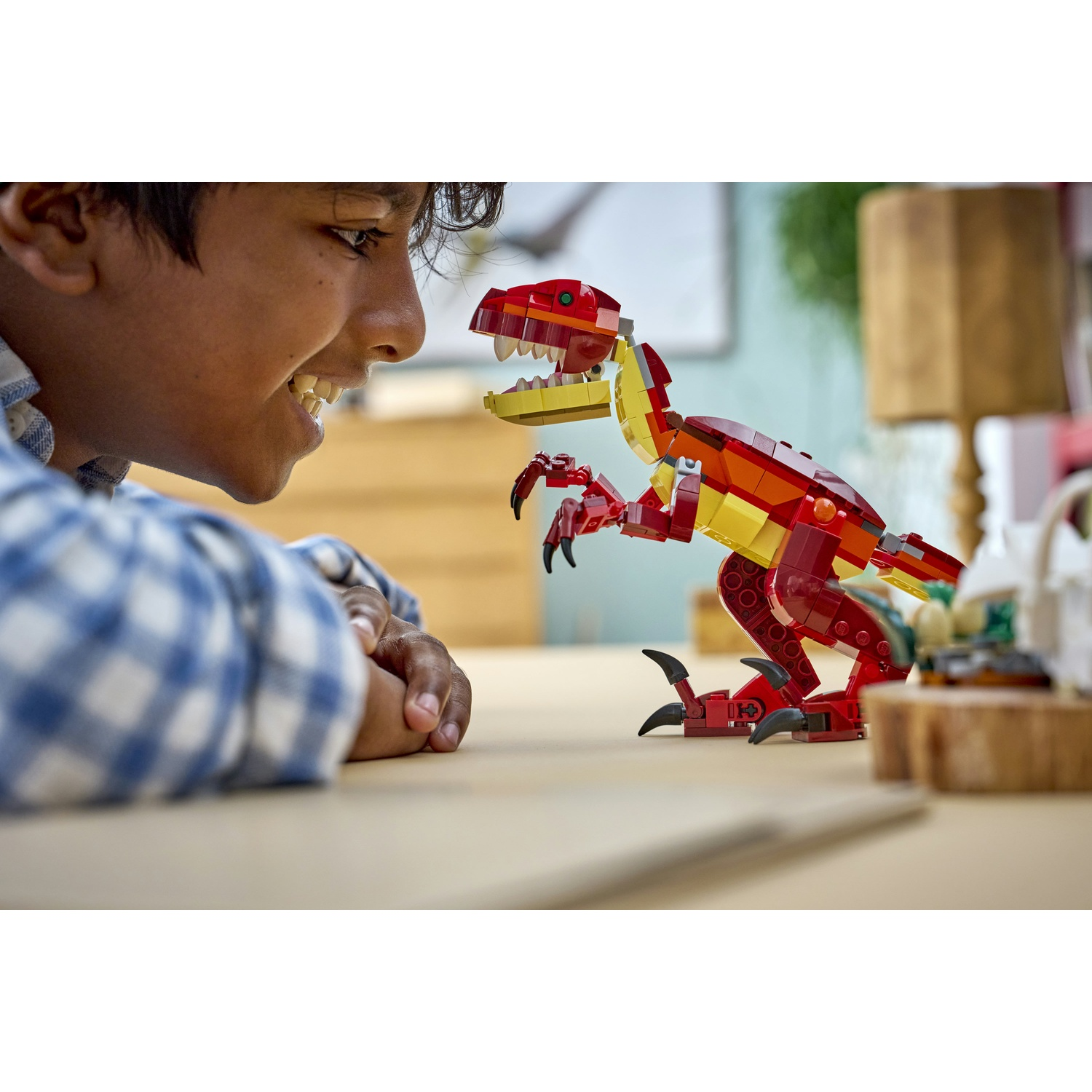 Un ragazzo sorride a un dinosauro Lego su un tavolo. Il dinosauro è rosso, giallo e arancione. Il ragazzo indossa una camicia a quadri blu.
