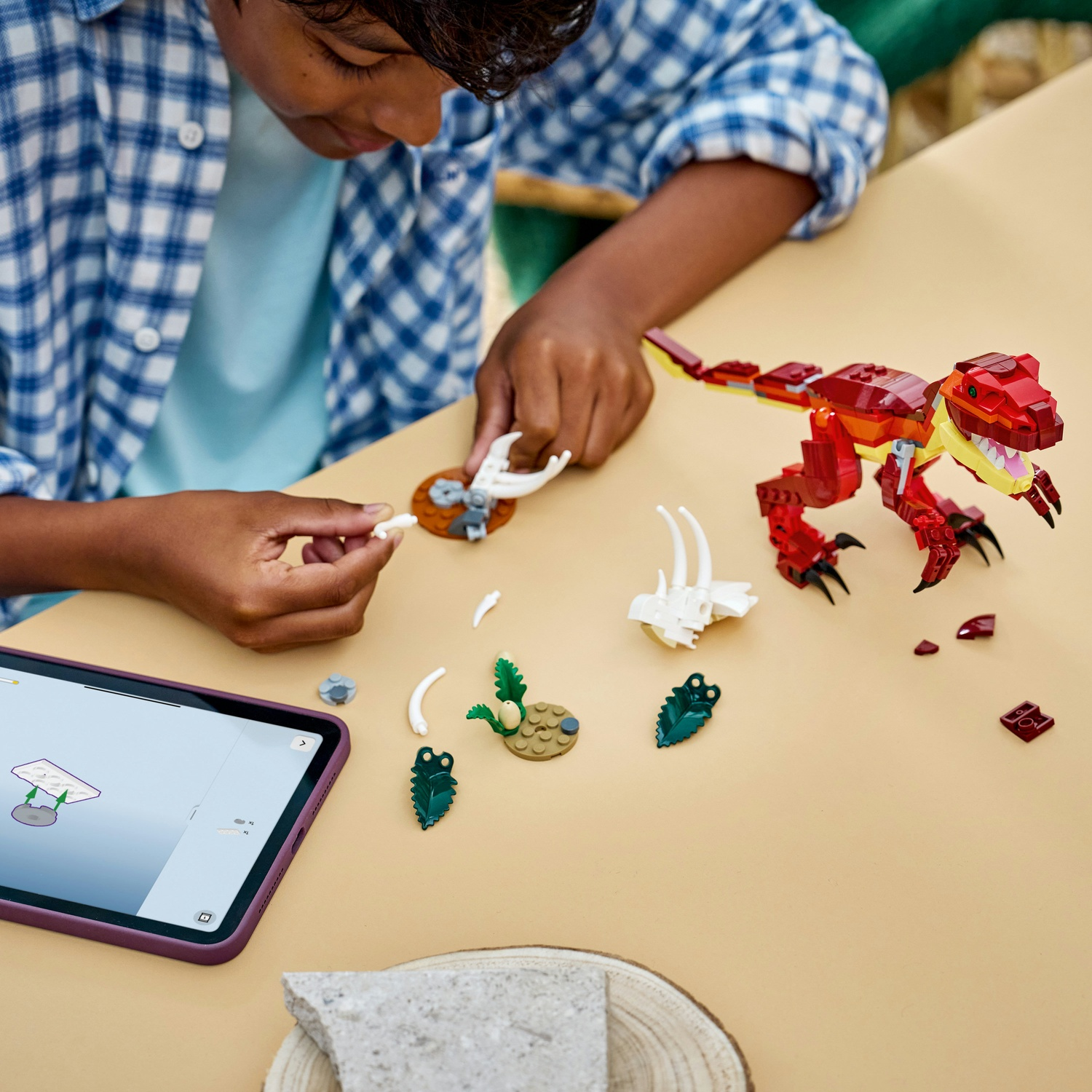 Un bambino assembla un giocattolo a forma di dinosauro rosso e giallo. Pezzi e tablet su un tavolo beige. Si vede una camicia a quadri blu.