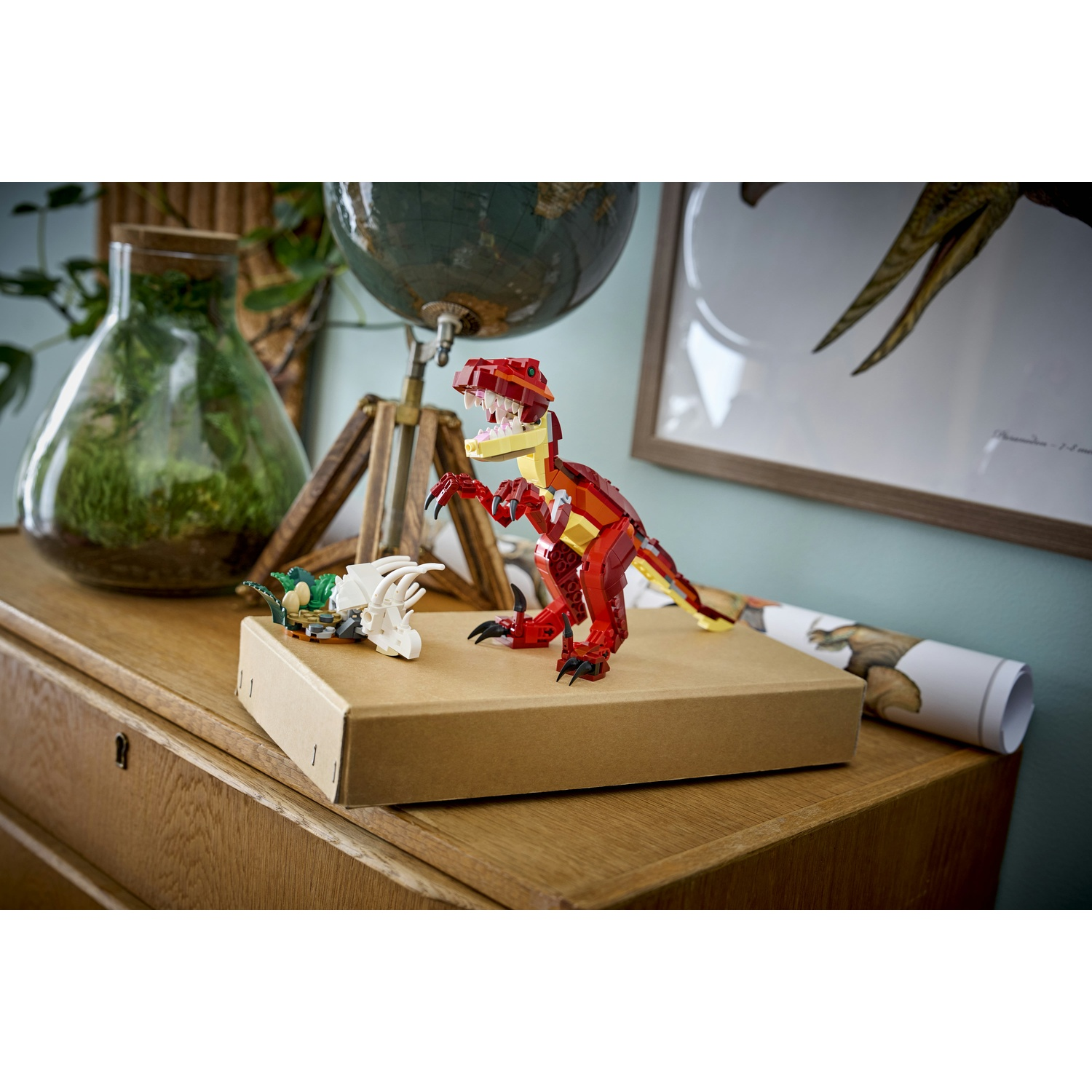 Un dinosauro Lego rosso e giallo è in piedi su una scatola di cartone. Un piccolo scheletro di dinosauro bianco e un'altra figura Lego verde sono vicini. Un globo è sullo sfondo.
