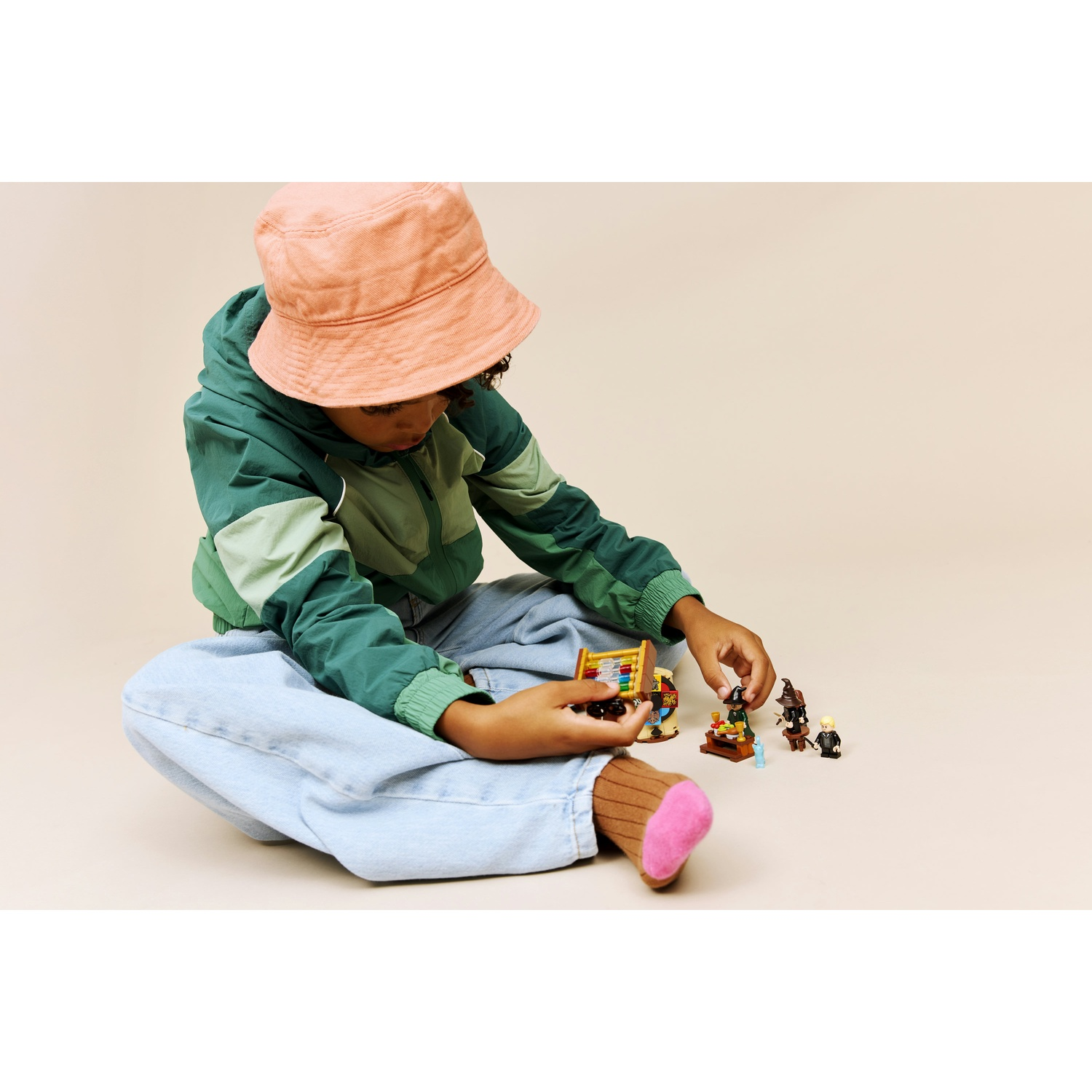 Un bambino con una giacca verde e un cappello rosa gioca con i personaggi Lego. Il bambino è seduto a gambe incrociate, concentrato sui giocattoli.