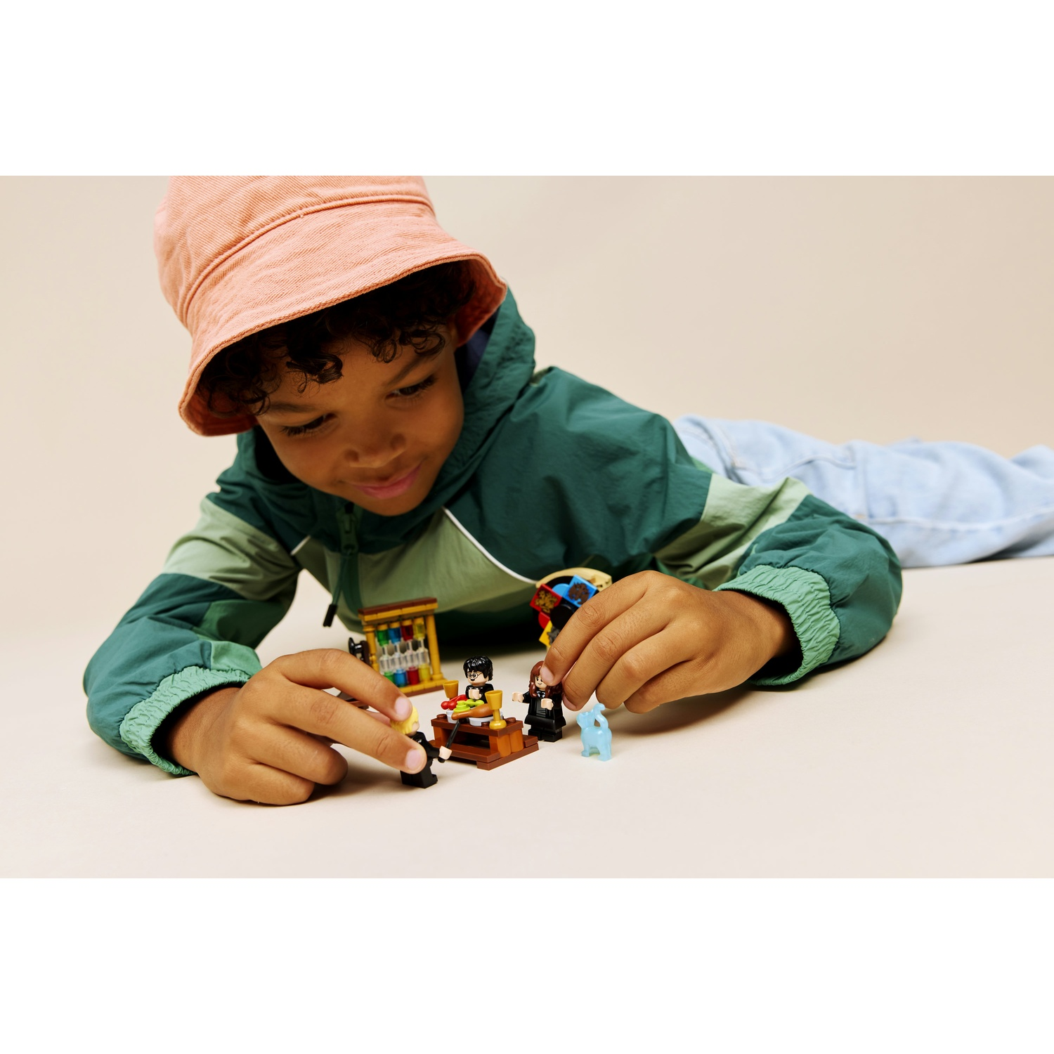 Un bambino con una giacca verde e un cappello rosa gioca con i personaggi Lego. Sono su una superficie beige con pezzi Lego.