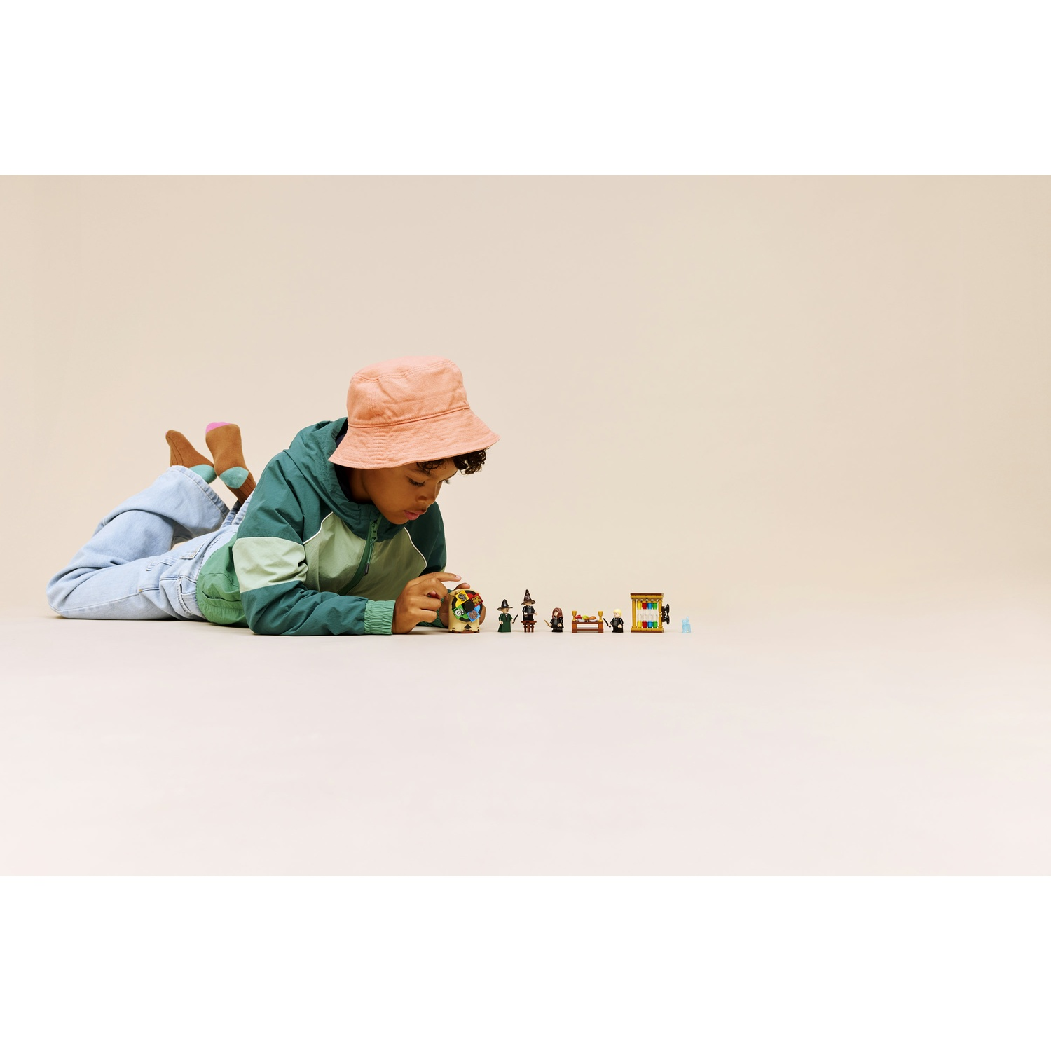 Un bambino con un cappello a secchio e una giacca verde è sdraiato sul pavimento, guardando piccole figure Lego. Lo sfondo è semplice.