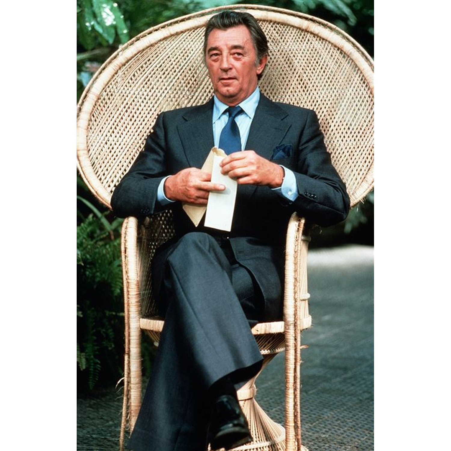Un homme en costume est assis dans une chaise en osier, tenant un papier. Le fond montre du feuillage vert.