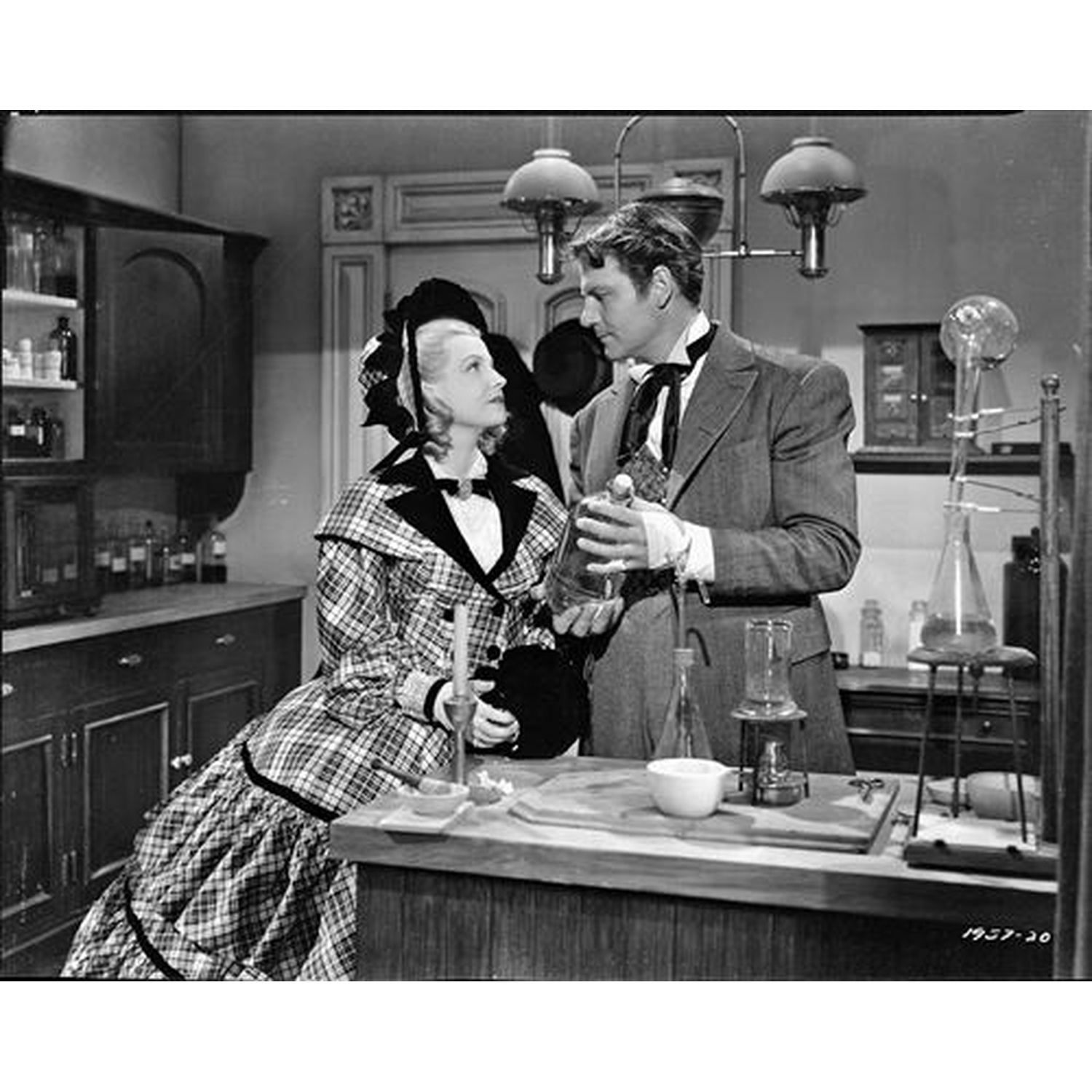 Een man en een vrouw in vintage kleding staan in een laboratorium. De man houdt een fles vast, de vrouw zit. Wetenschappelijke apparatuur is zichtbaar.