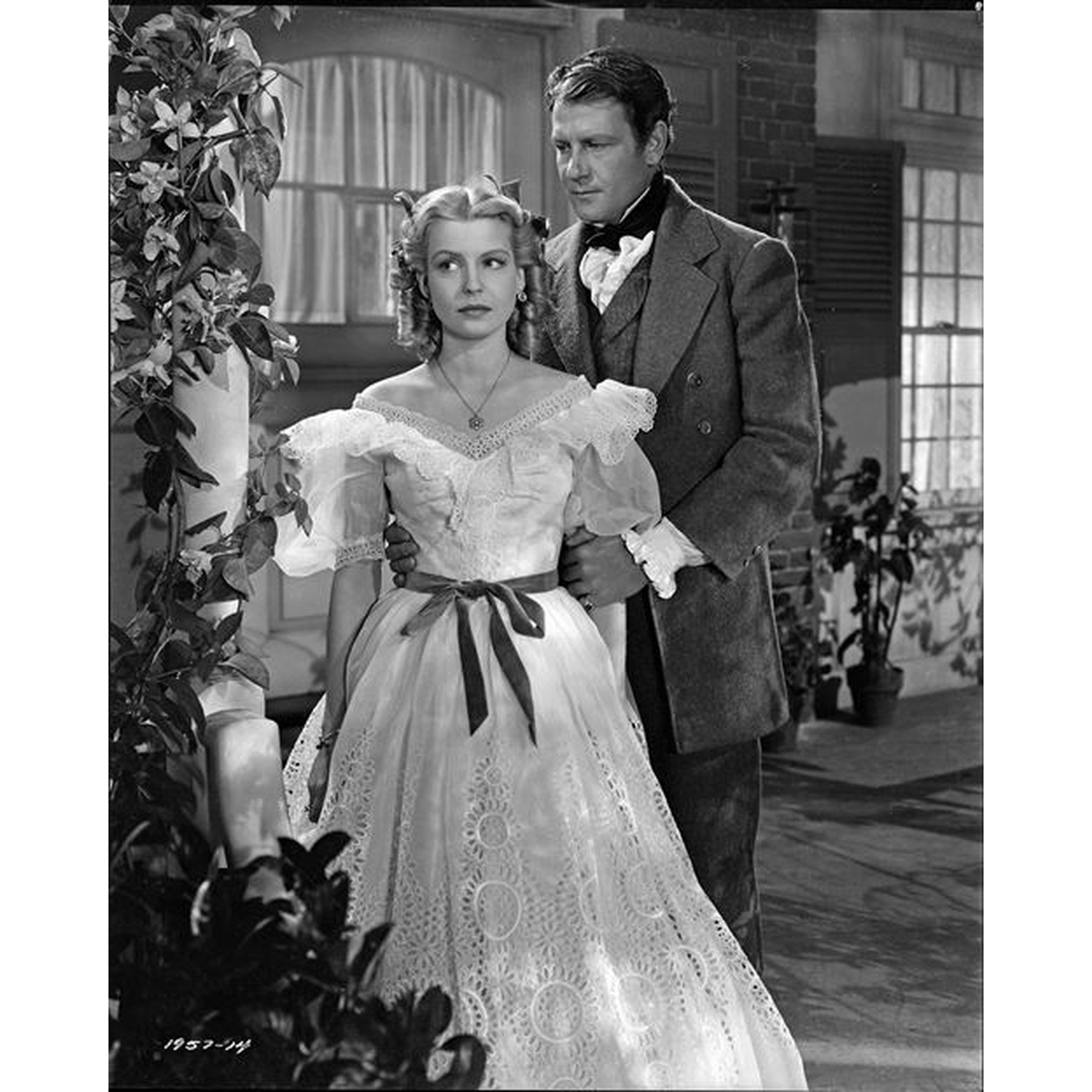 Un homme et une femme se tiennent près. Elle porte une robe blanche, lui un costume. Ils sont près d'un bâtiment avec des fenêtres.