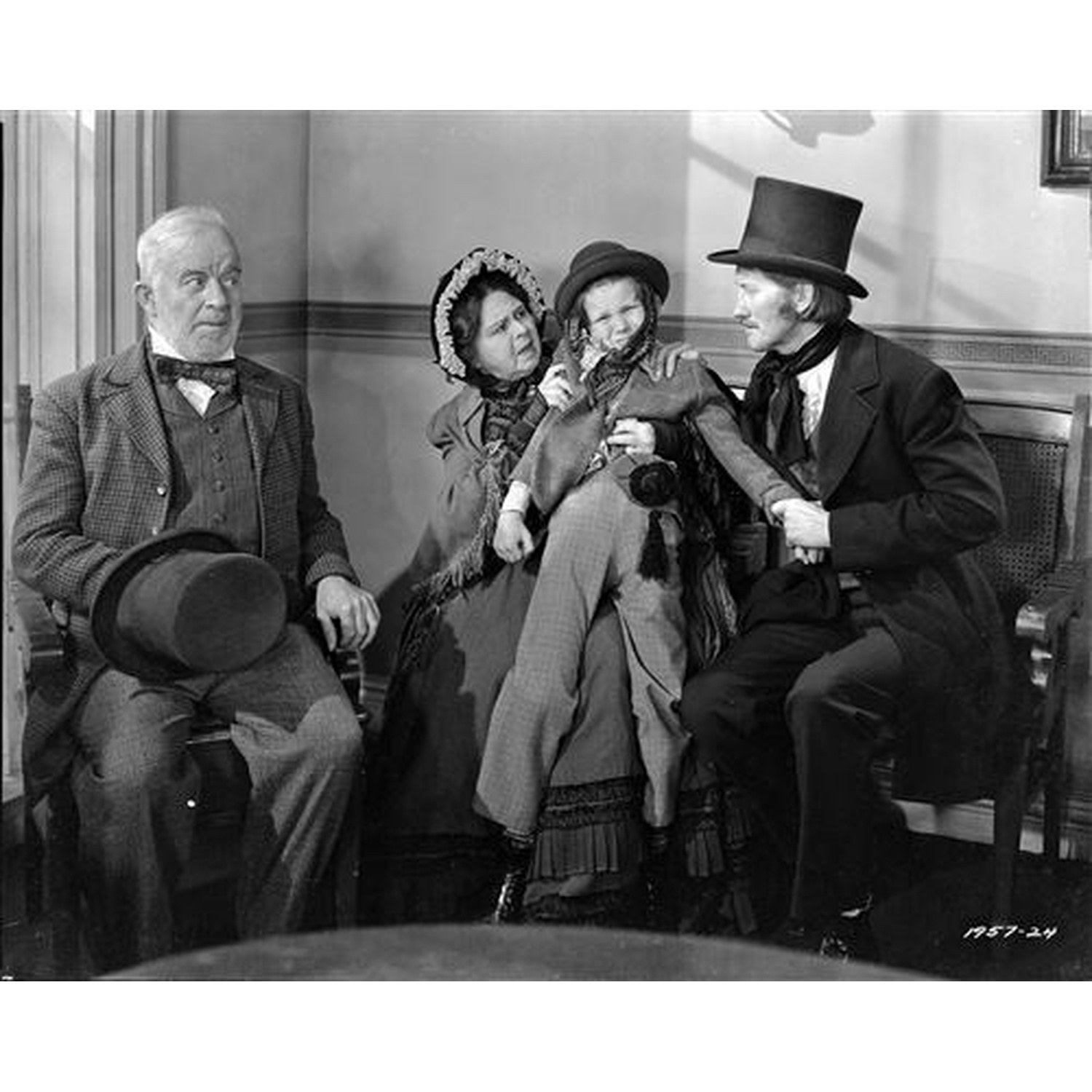 Quatre personnes en tenue d'époque, assises. Un homme en haut-de-forme tient un enfant. D'autres regardent. Noir et blanc, intérieur.