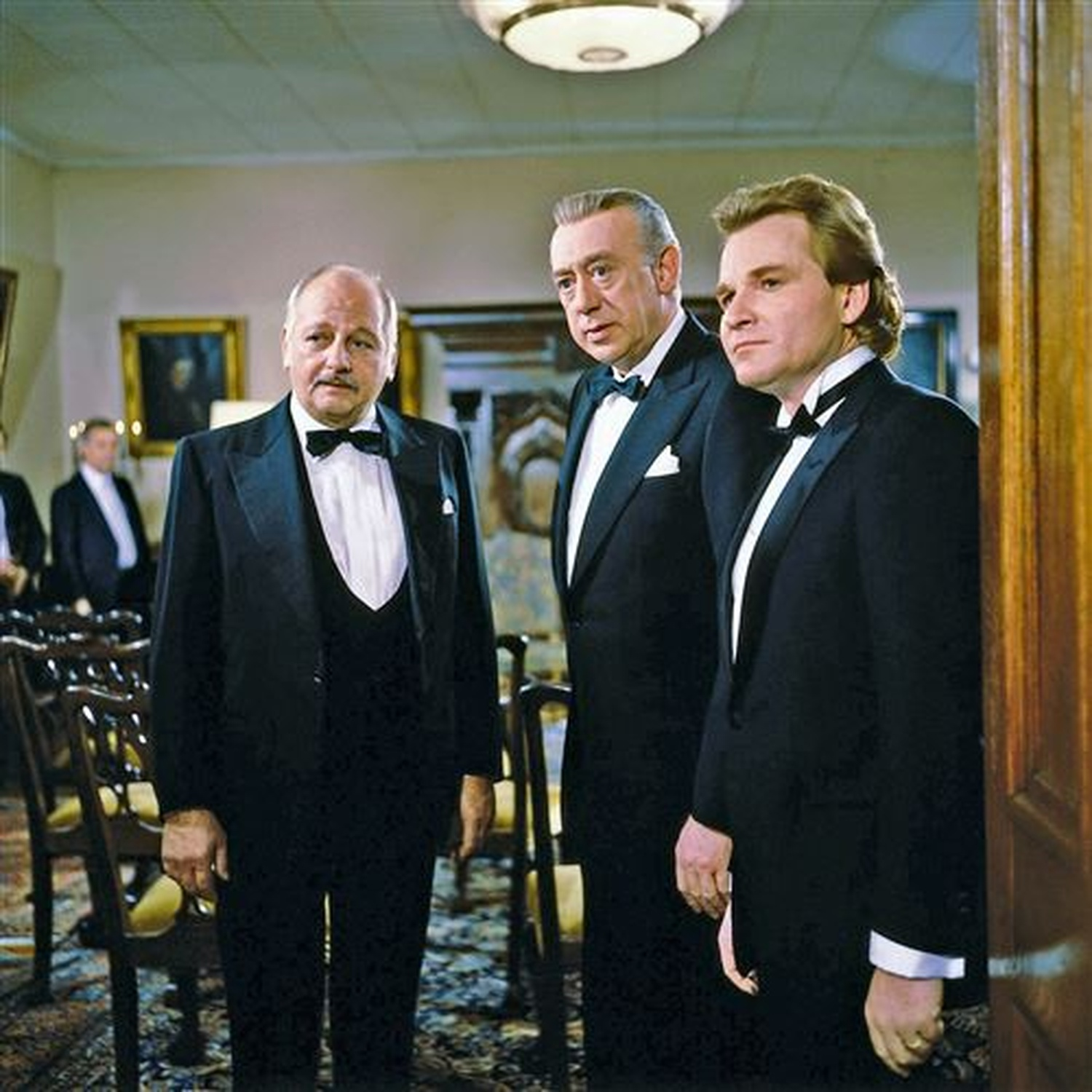 Three men in tuxedos stand indoors. The man in front has a mustache. Others are in the background. Wooden chairs and a door are present.