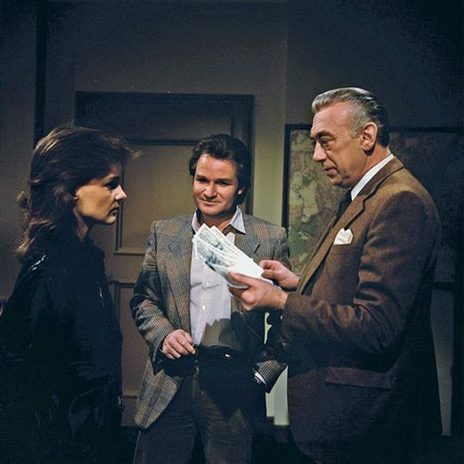 Three people stand indoors. A man holds money, facing two others. A woman in black looks on. Background includes a door and a map.