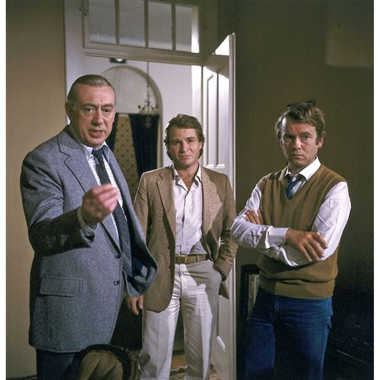 Three men in suits stand in a room. One points, another has arms crossed, the third has hands in pockets. Neutral background.