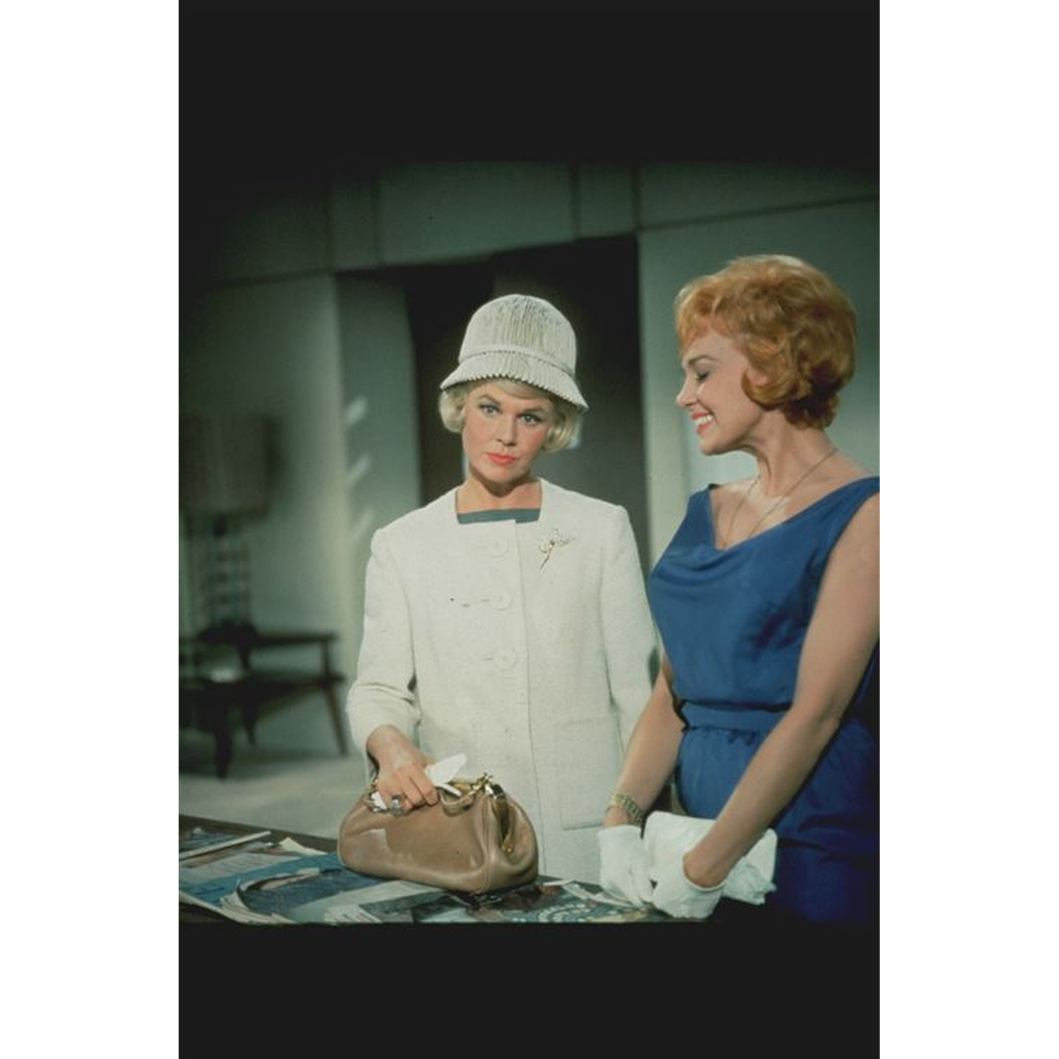 Deux femmes en tenue des années 1960. Une en costume et chapeau blancs, l'autre en robe bleue. Elles sont à une table, souriantes.