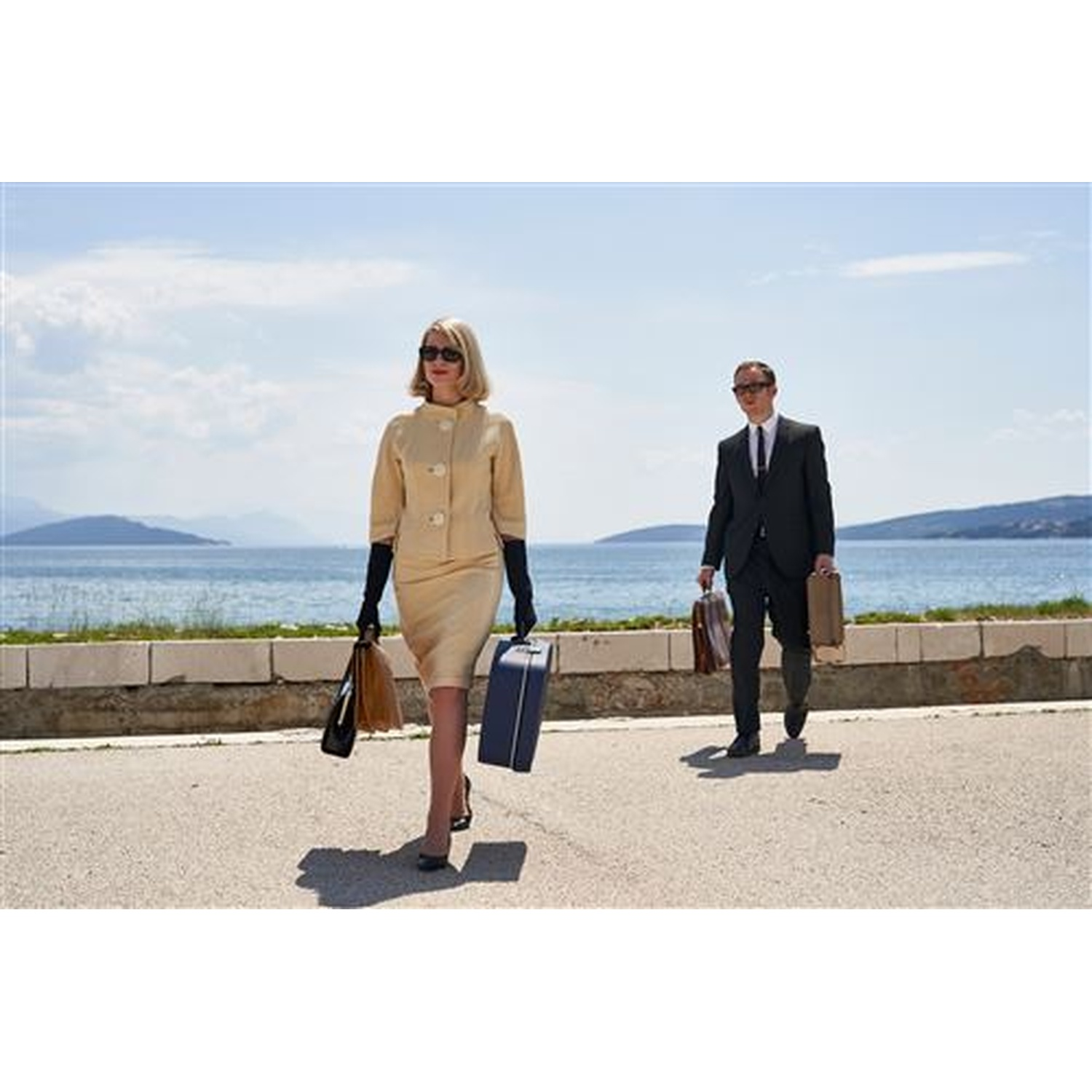A woman in a cream suit and a man in a black suit walk on a road with suitcases. The sea and mountains are in the background.