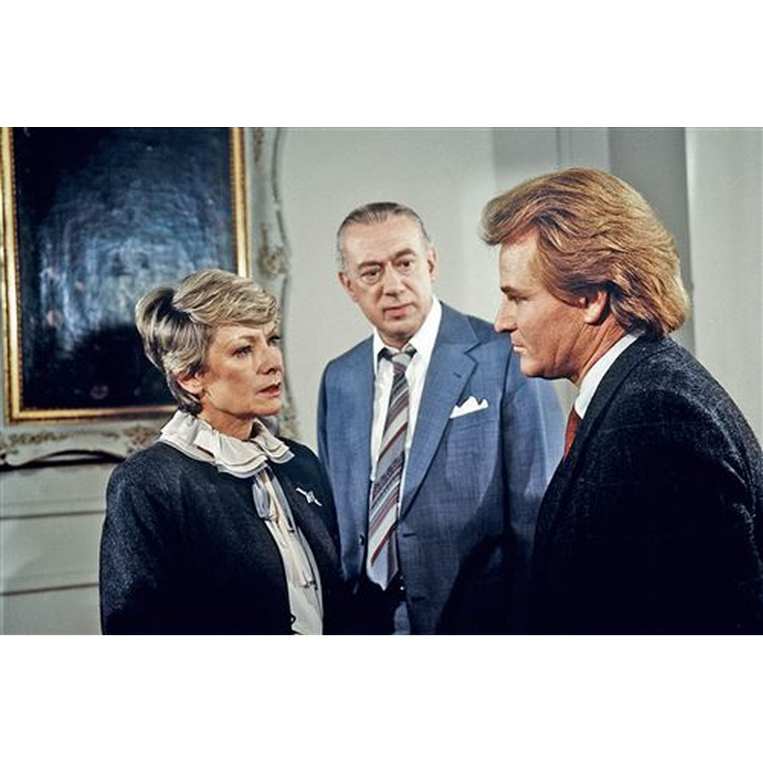 Three people in suits are in a room. A woman looks to the left, a man looks forward, and another man looks right.