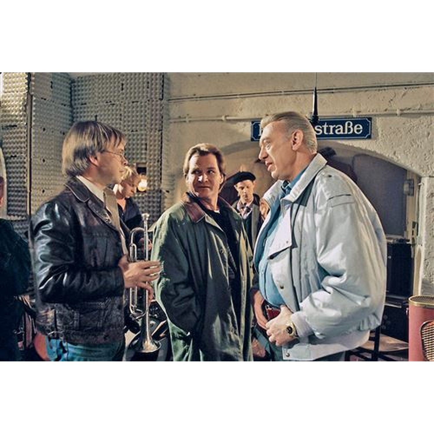 Men in jackets talking near a street sign. One holds a trumpet. Background includes a wall, a door, and other people.