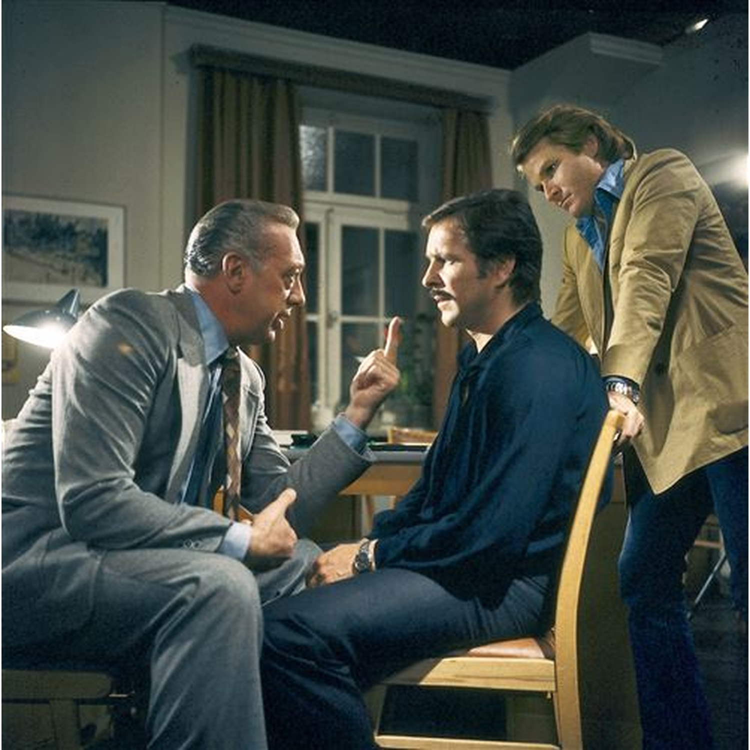 Three men in a room. One points a finger. Another sits, and a third stands behind the seated man, holding the chair.