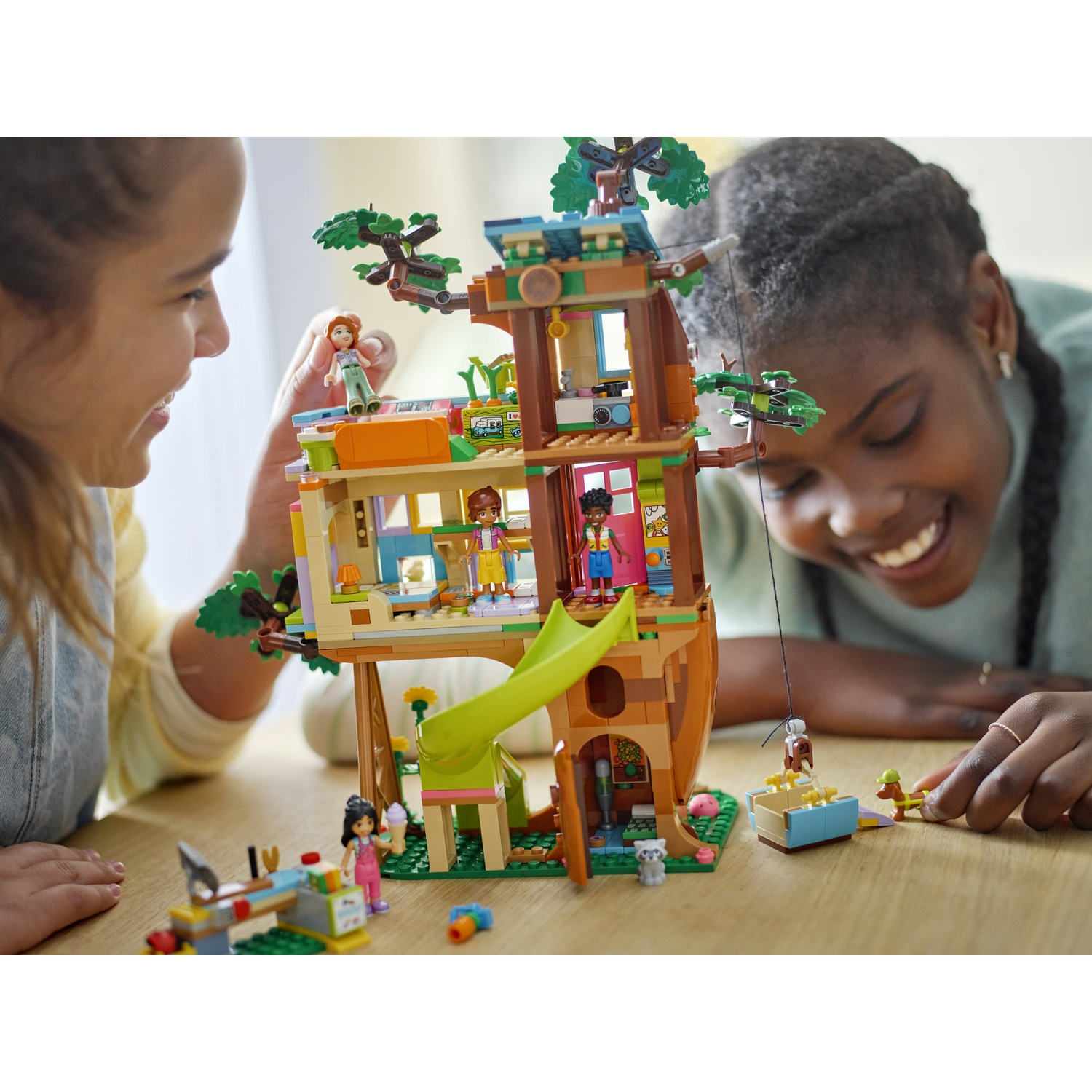Due ragazze giocano con una casa sull'albero Lego. La casa sull'albero ha uno scivolo e diverse figure Lego. Volti sorridenti e atmosfera felice.