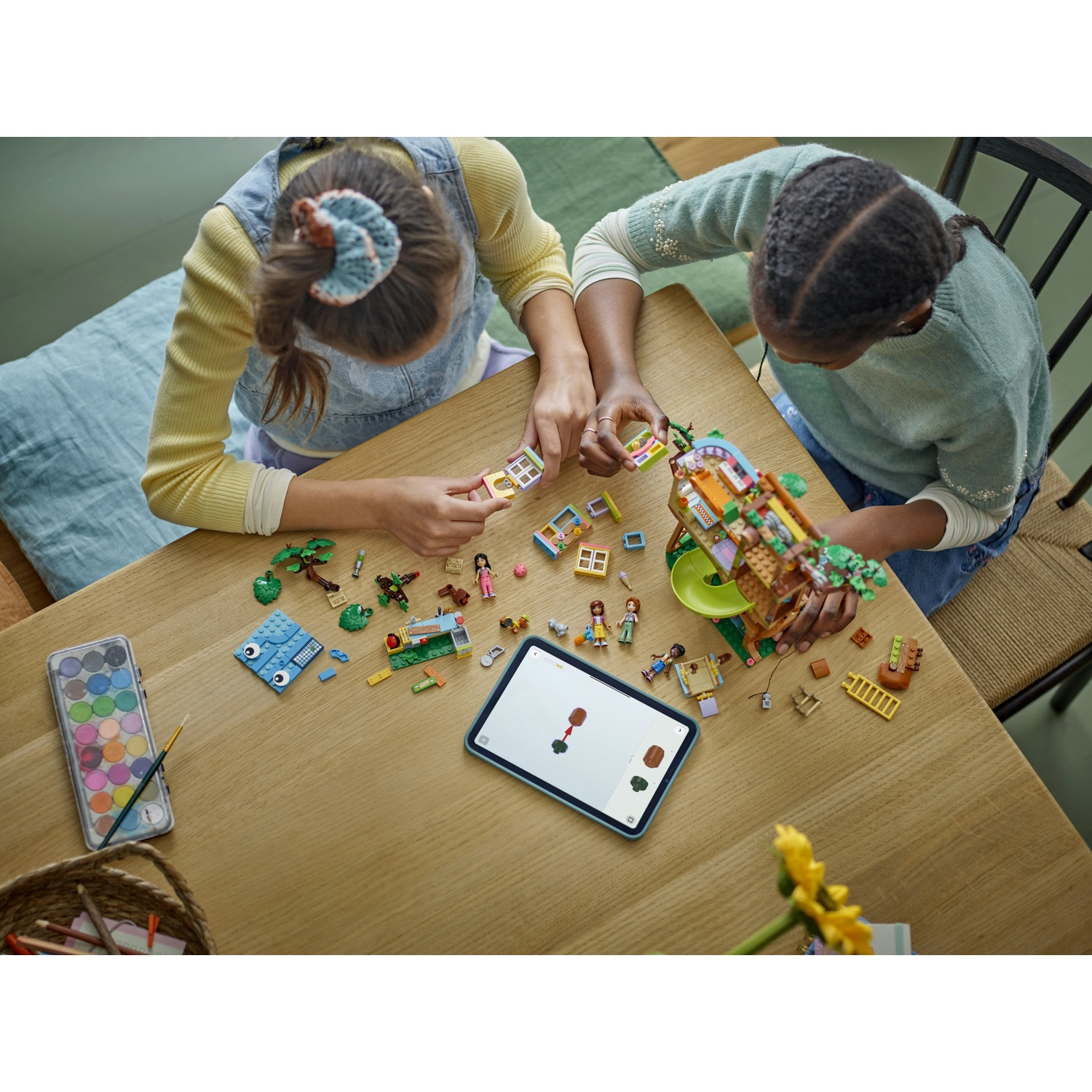 Due bambini costruiscono Lego su un tavolo di legno. Un tablet è nelle vicinanze. Vernice e matite sono visibili. I pezzi Lego sono sparsi.