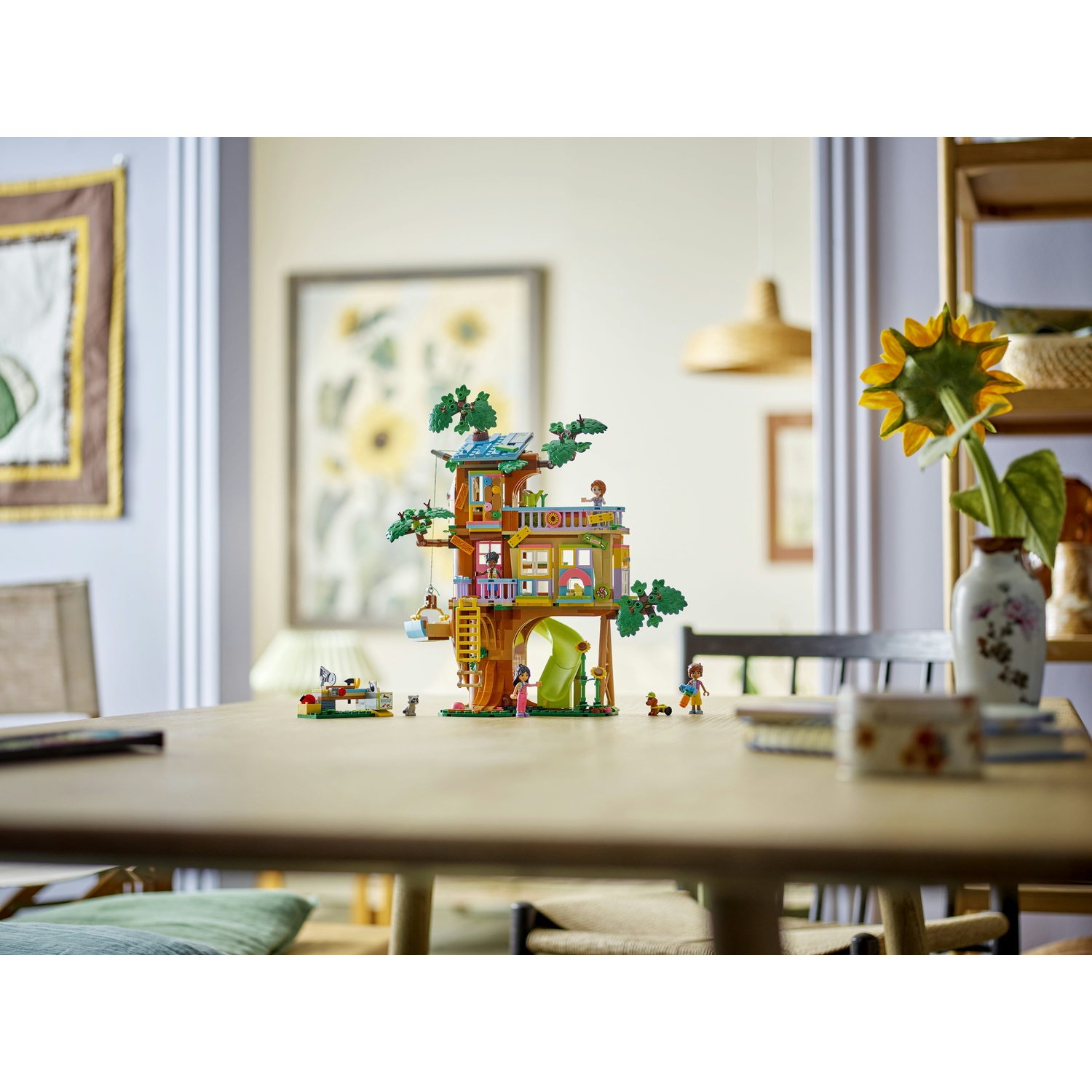 Una casa sull'albero Lego su un tavolo. Include figure, uno scivolo e un'altalena. Un girasole è in un vaso.