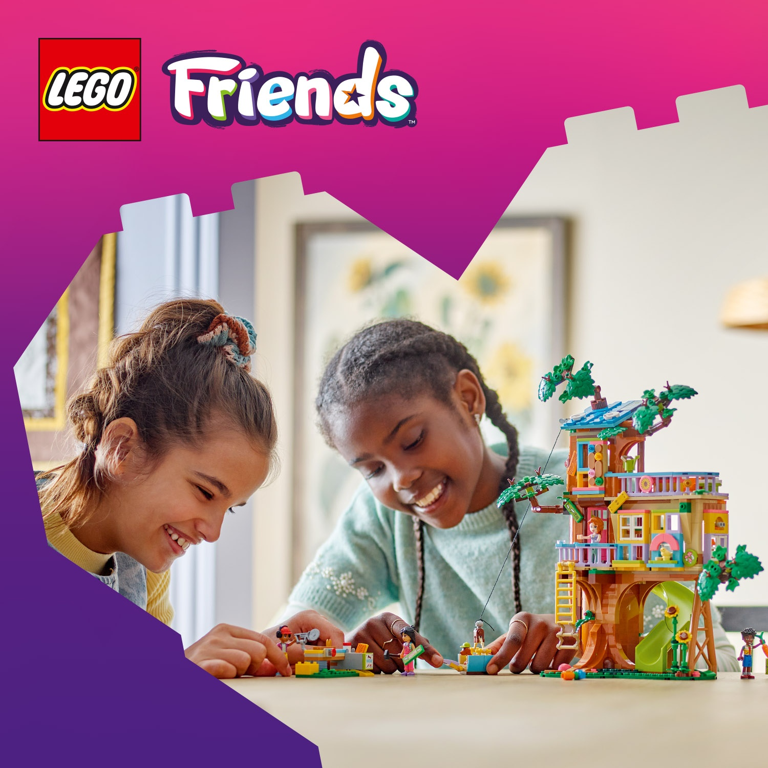 Due ragazze sorridono mentre costruiscono una casa sull'albero LEGO Friends. La casa sull'albero è marrone con scivoli e balconi verdi.