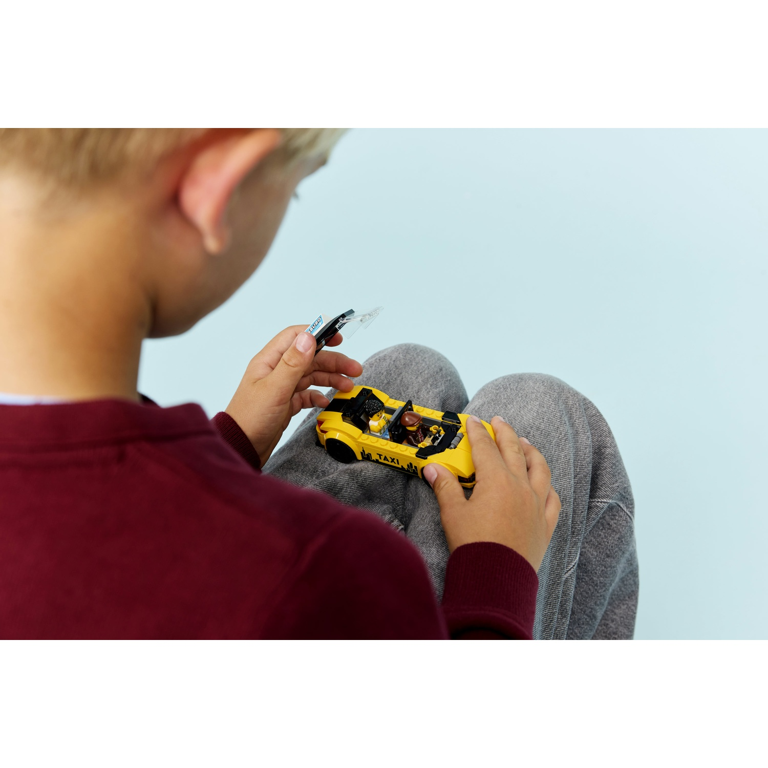 Un bambino tiene in mano un taxi giocattolo giallo. Il bambino indossa una camicia bordeaux e pantaloni grigi. Lo sfondo è azzurro.