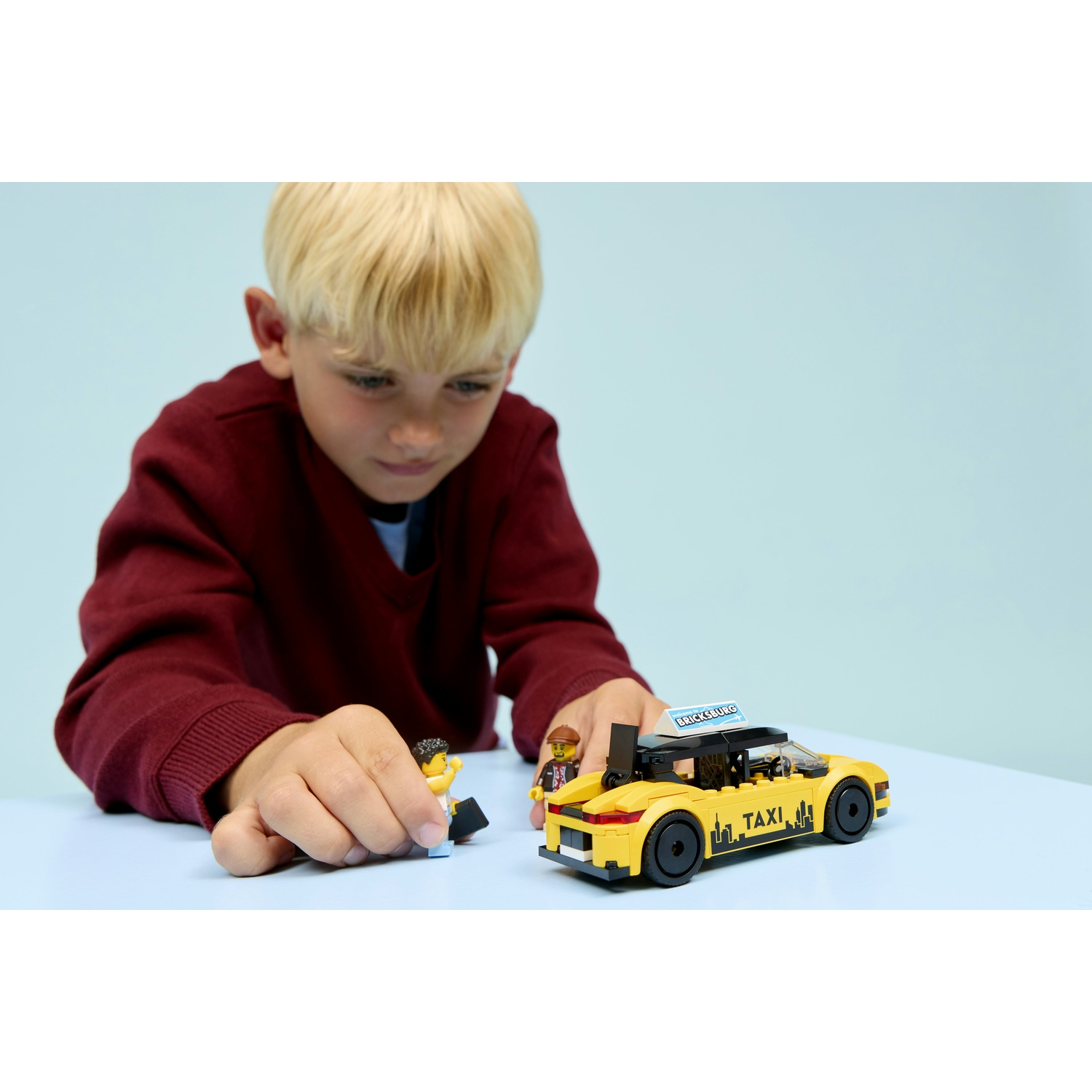 Un ragazzo con un maglione rosso gioca con un taxi giocattolo giallo e due personaggi Lego su una superficie azzurra.