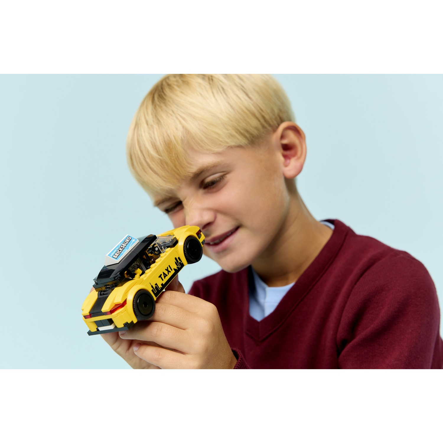 Un ragazzo tiene in mano un taxi Lego giallo. Il taxi ha strisce nere e la scritta TAXI. Il ragazzo indossa un maglione bordeaux e sorride.