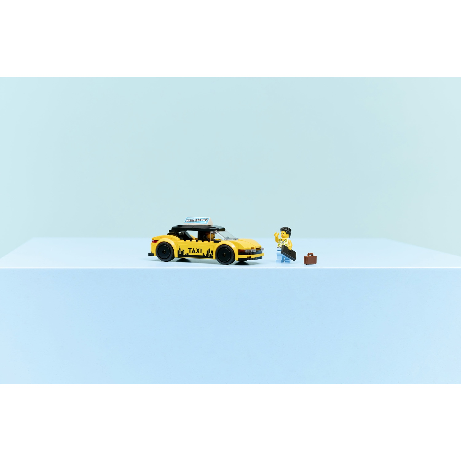 Un taxi giallo con un cartello, un personaggio Lego con una valigia e un'altra piccola valigia. Tutto su una superficie blu.