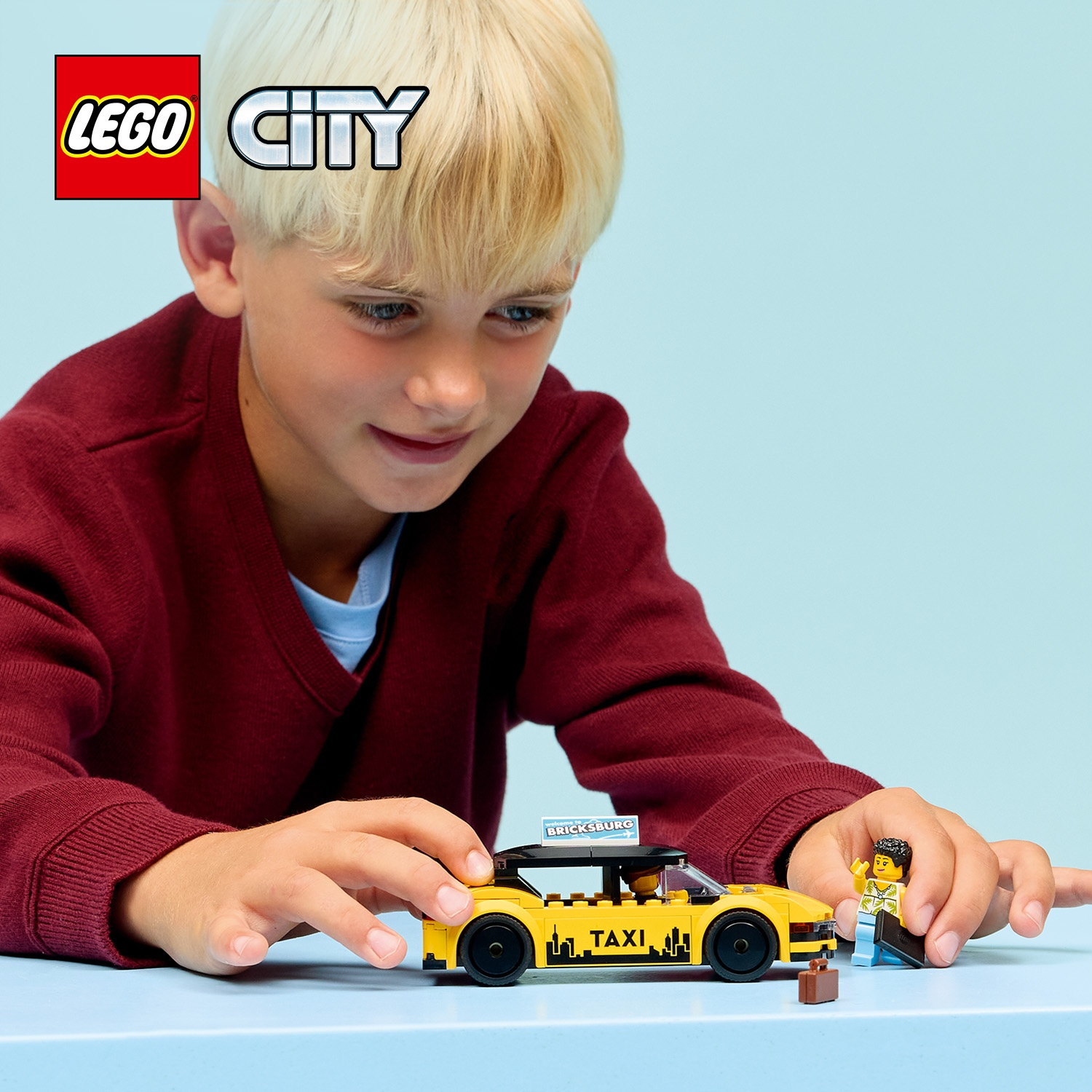 Un ragazzo gioca con un taxi LEGO giallo. C'è un conducente e un passeggero con una valigia. Il ragazzo indossa un maglione rosso.